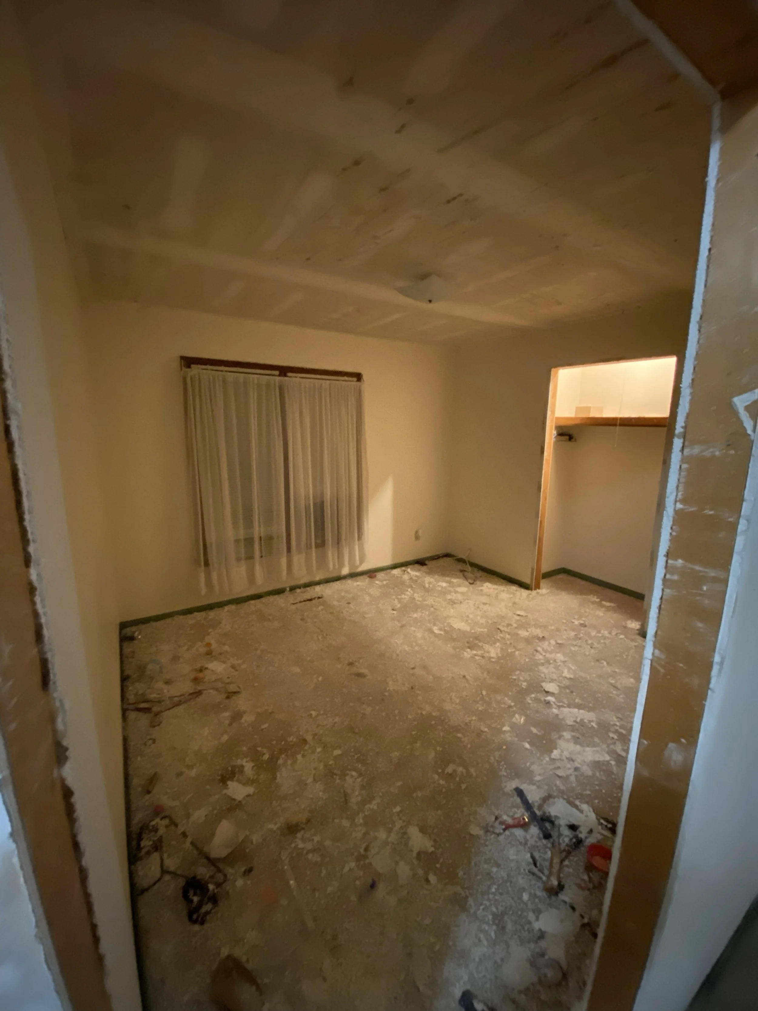 Interior of a room under renovation with a window with curtains, a small closet space, and a floor covered in debris and construction materials.