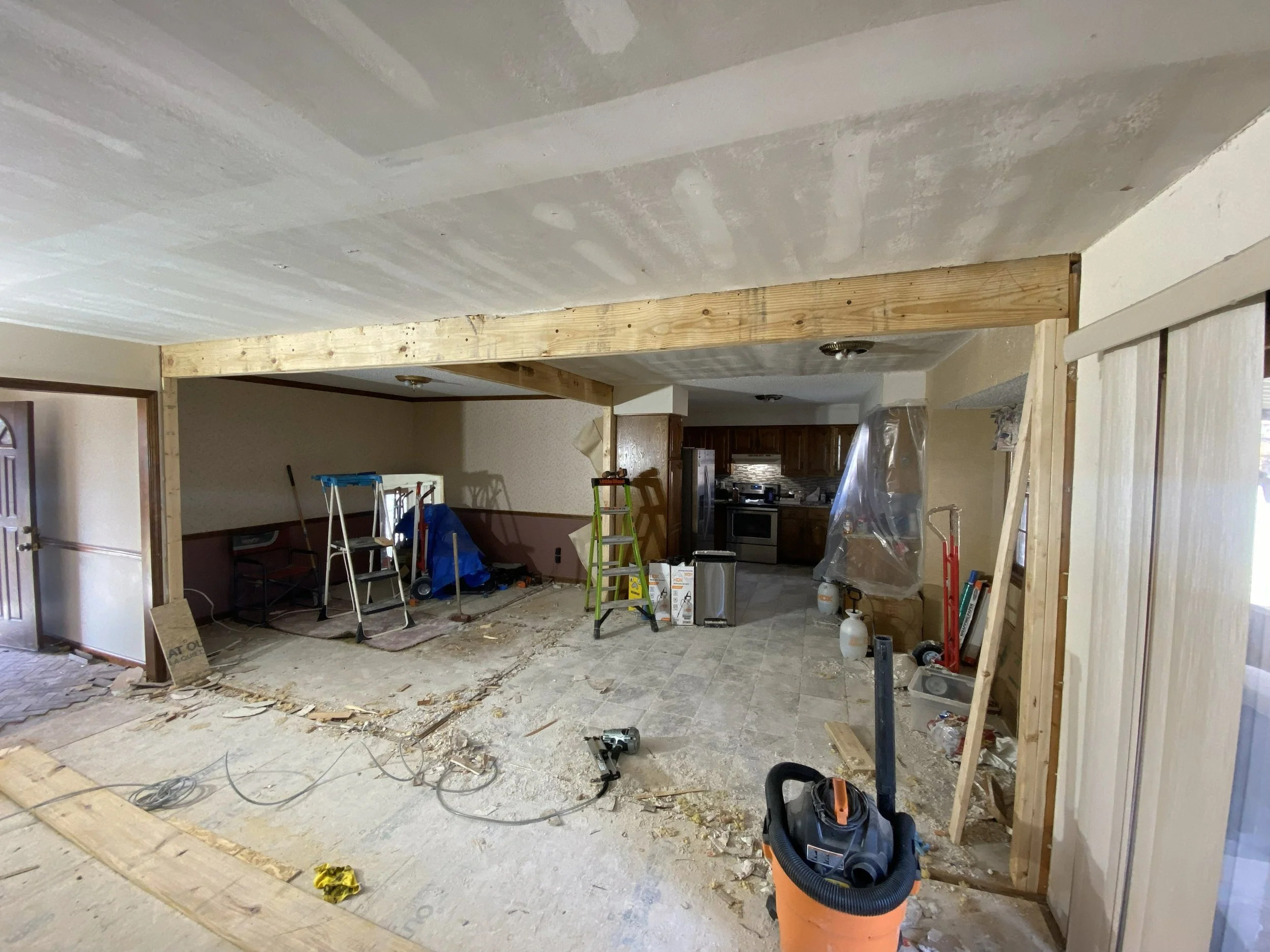 Living room under renovation with exposed wooden framing, construction tools, and off-center kitchen visible in background.