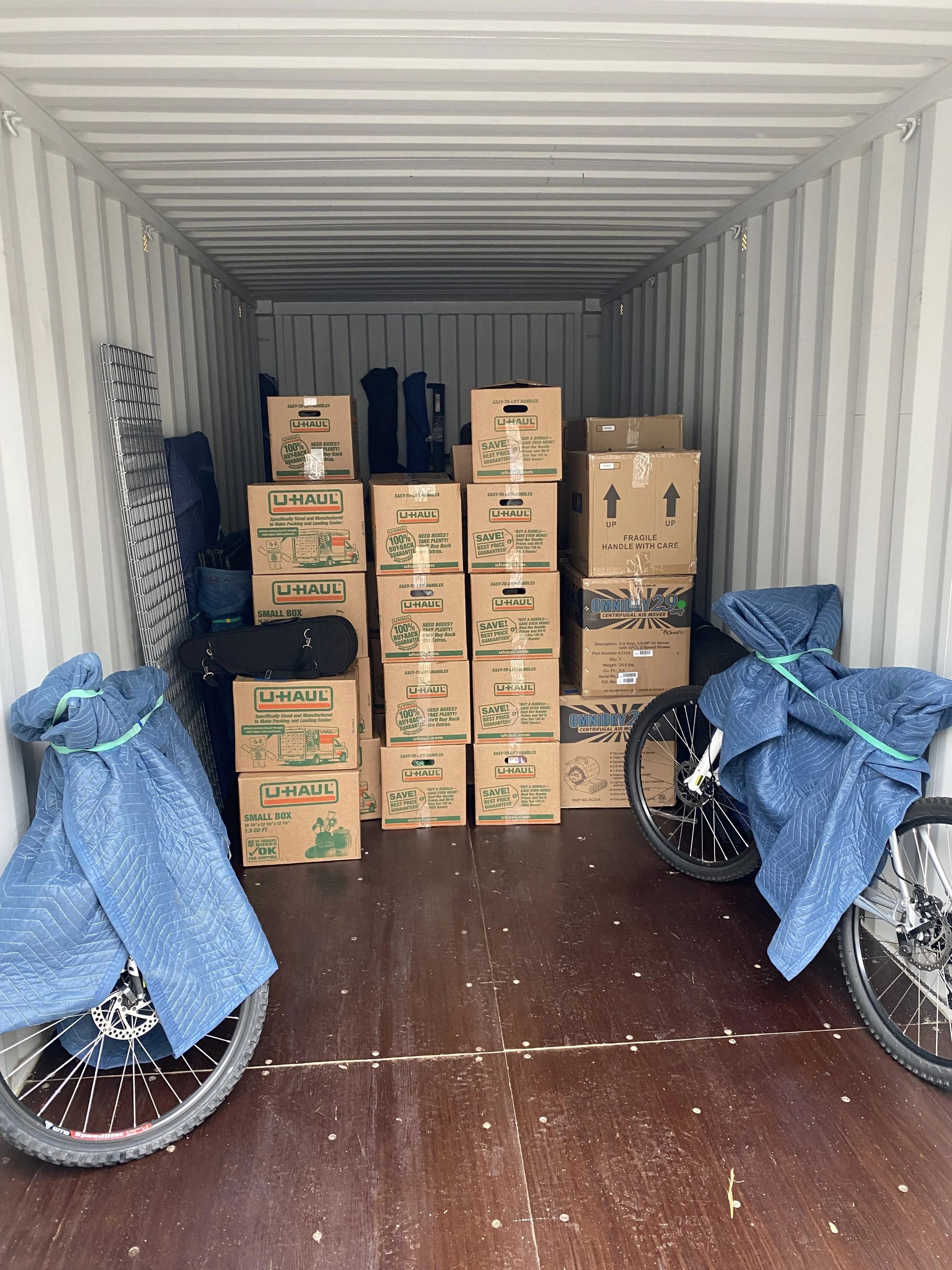 Inside a storage unit filled with packed boxes, bicycles with blue covers, and various stored items.