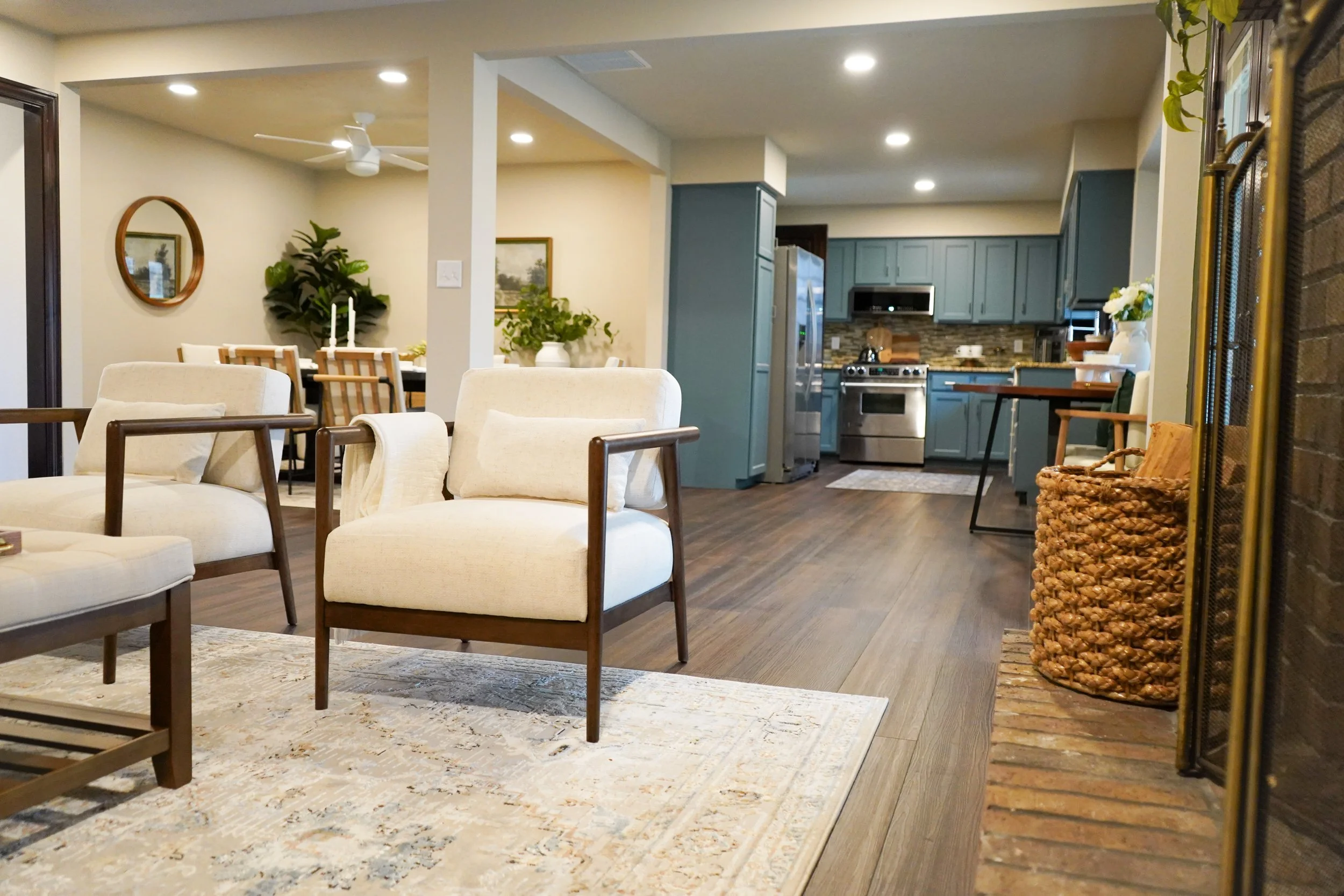 Interior of a modern living room with off-white armchairs, a Rug, a kitchen area with blue cabinets, stainless steel appliances, dining table with chairs, fireplace with a screen, and decorative plants