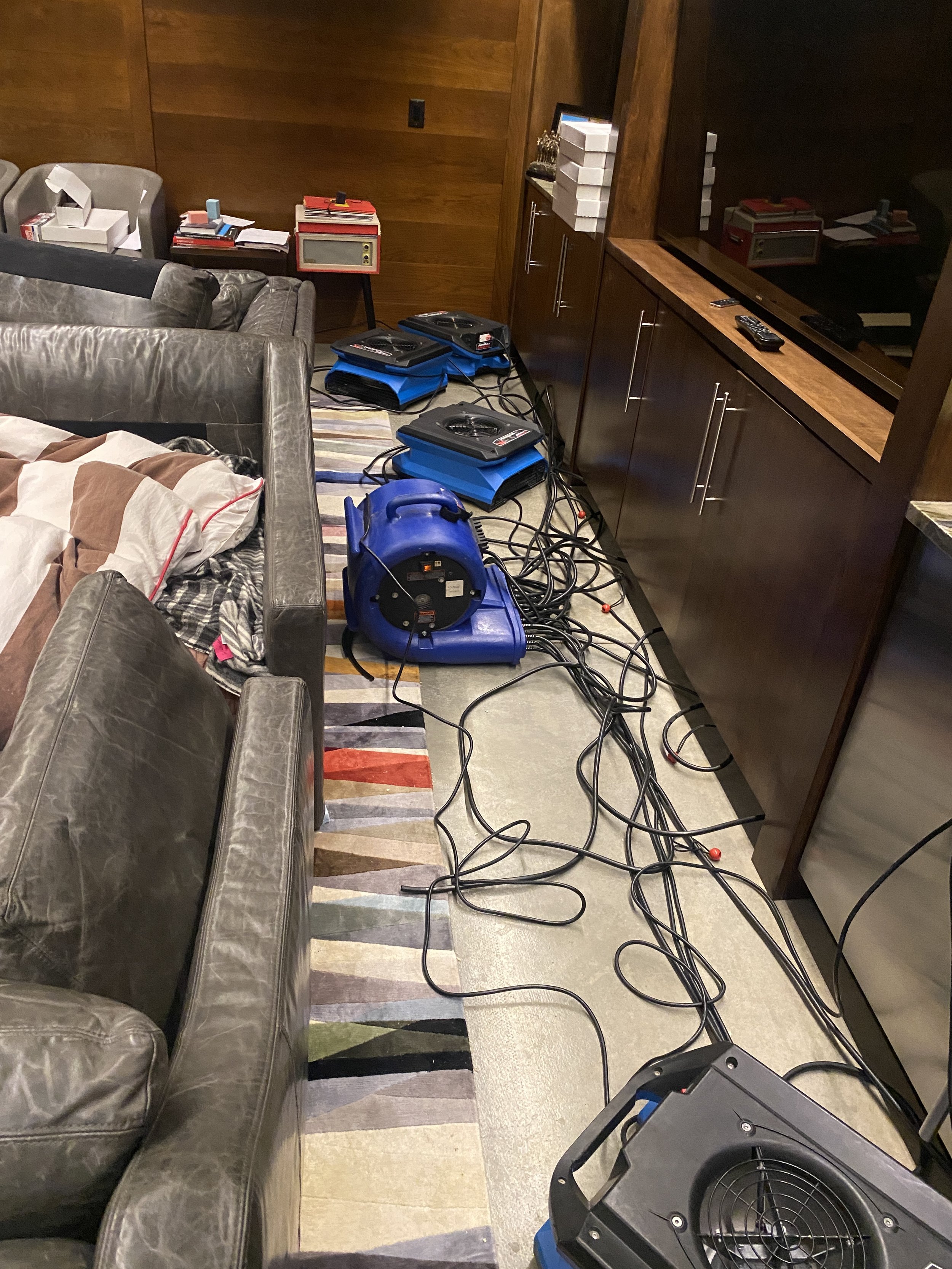 Tools and equipment set up on a carpeted floor with tangled cords, a blue inflatable air mover, and plastic containers in a room with brown wood-paneled walls, a leather sofa, and a cabinet.