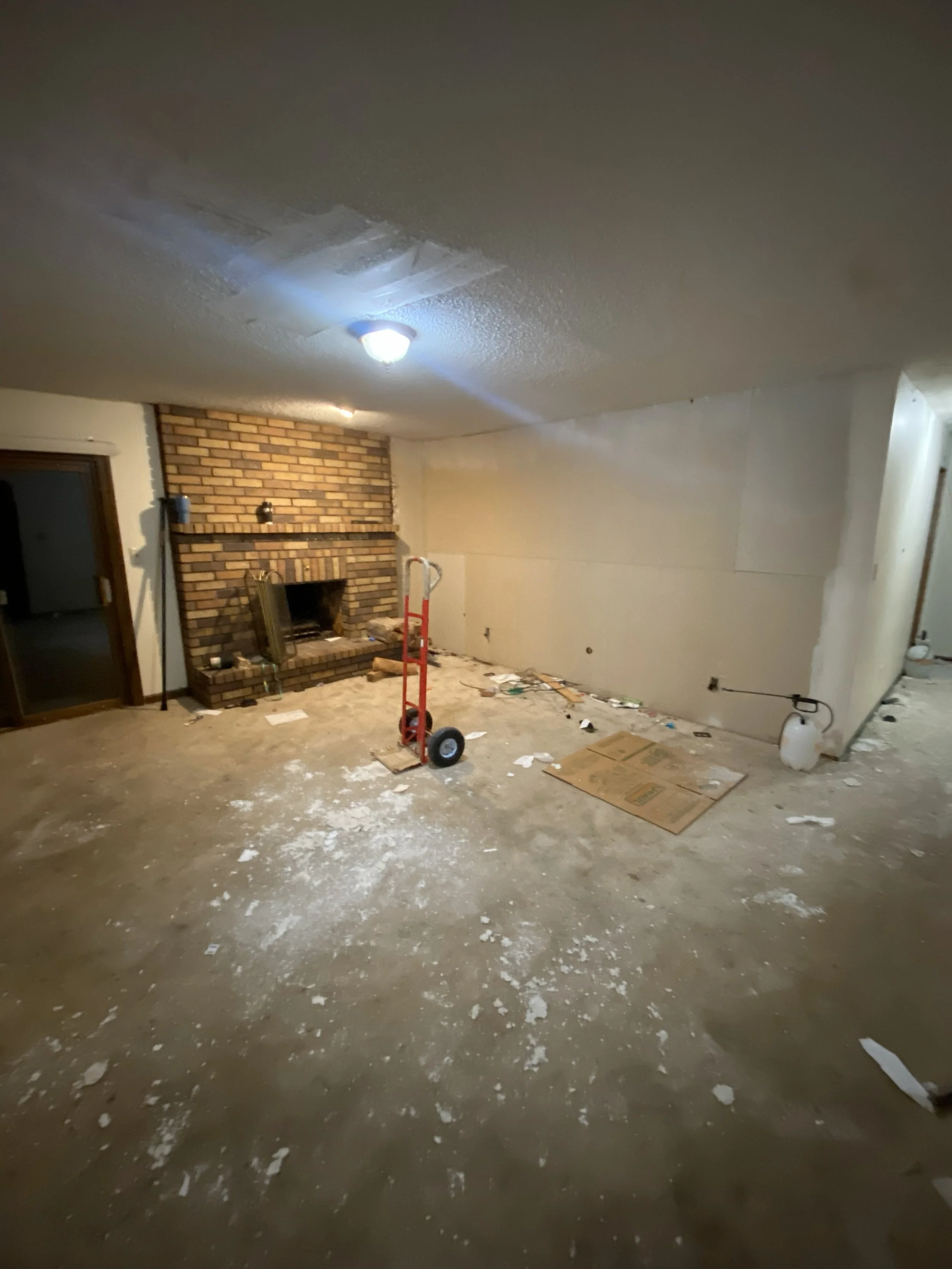 Empty room undergoing renovation with a brick fireplace, scattered debris, a hand truck, and construction tools.