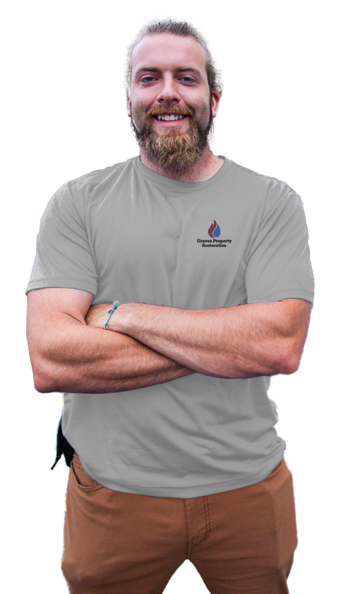 A smiling man with a beard and long hair tied back, arms crossed, wearing a gray t-shirt with a logo that reads "Groves Property Restoration" and brown pants.