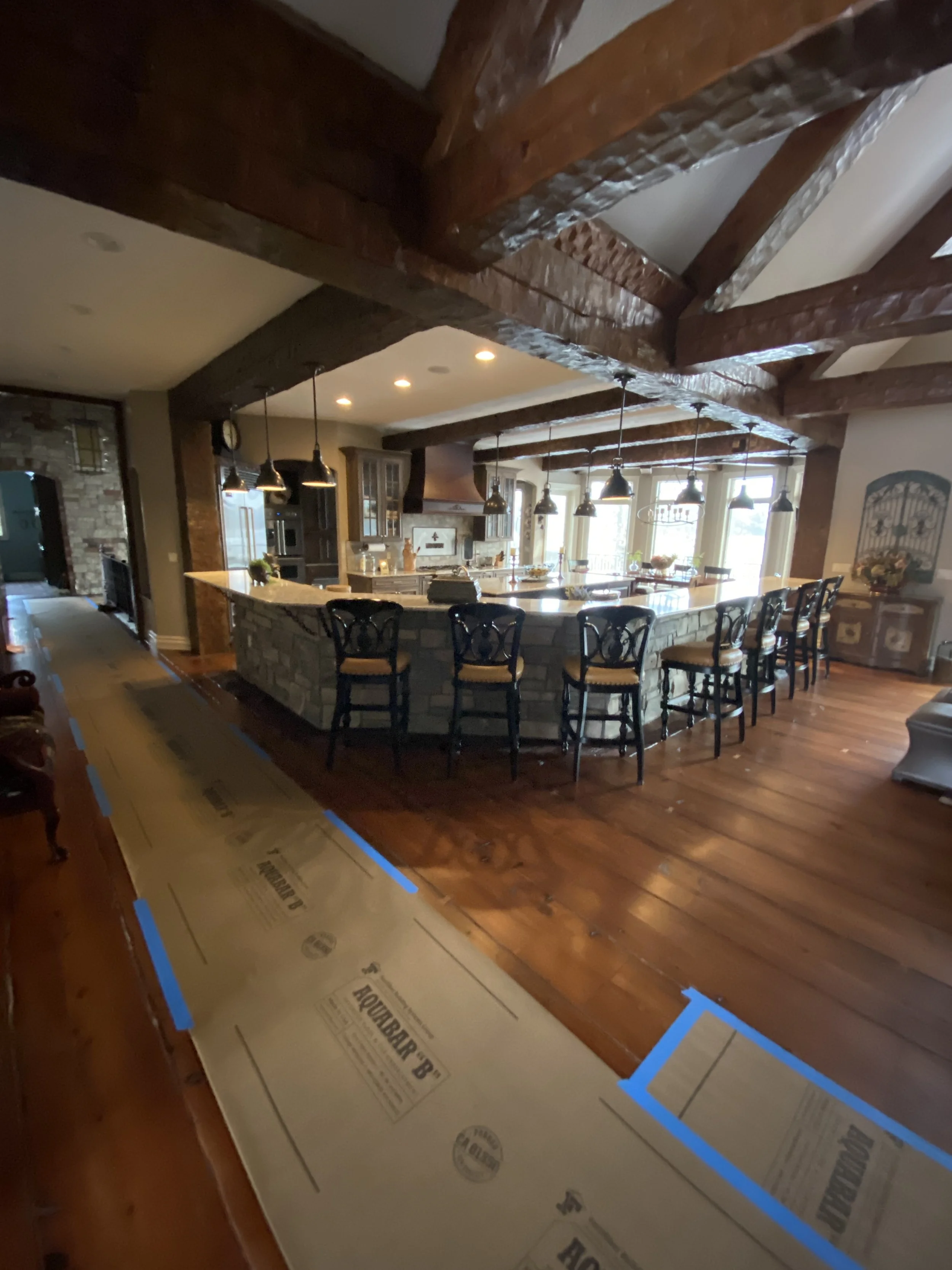 Interior of a kitchen and dining area with exposed wooden beams, a large island with pendant lights, and hardwood flooring. Blue painter's tape on the floor indicates ongoing renovation.