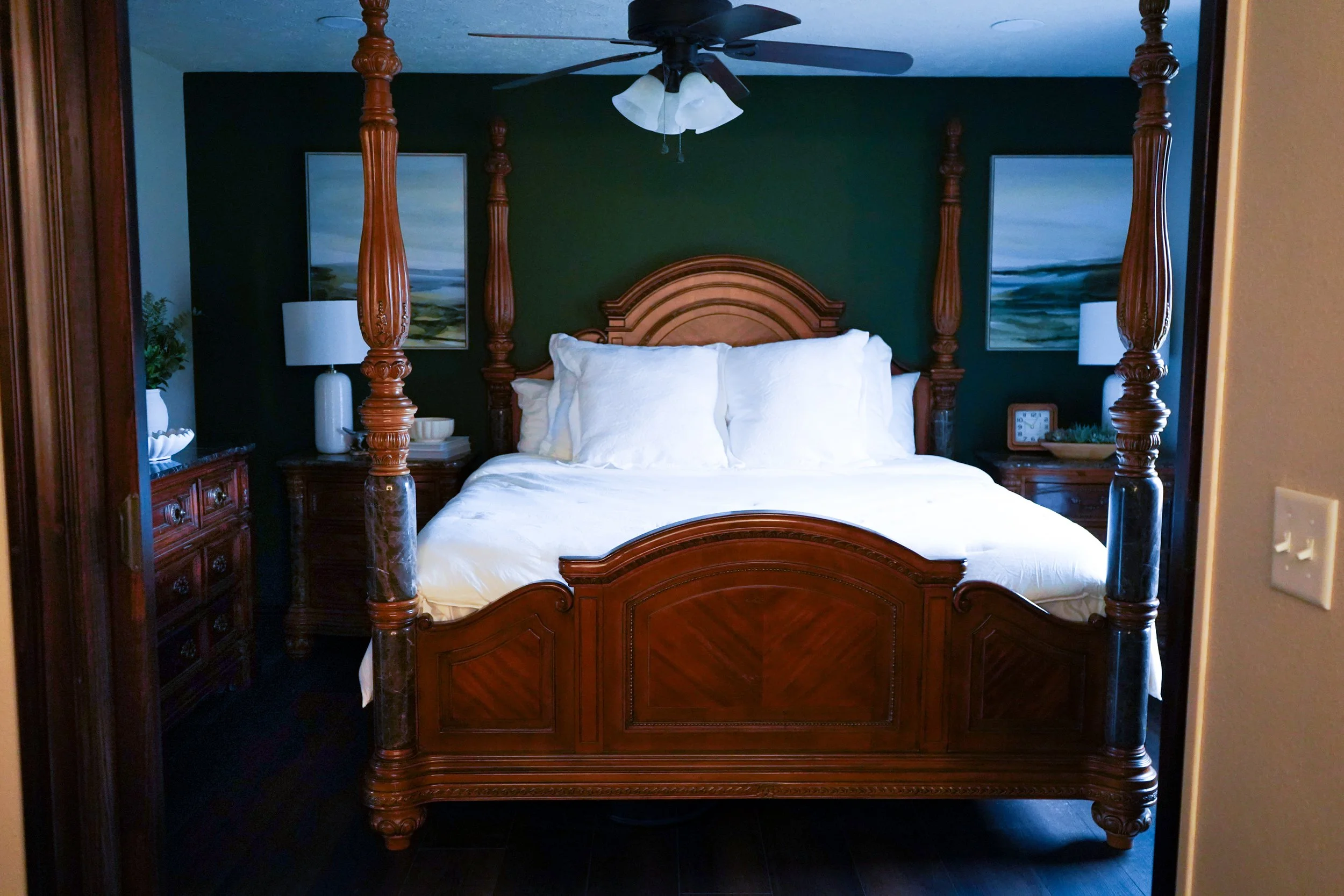 A bedroom with a four-poster wooden bed with white bedding and pillows, lamp on bedside tables, paintings on dark green wall, ceiling fan with light.
