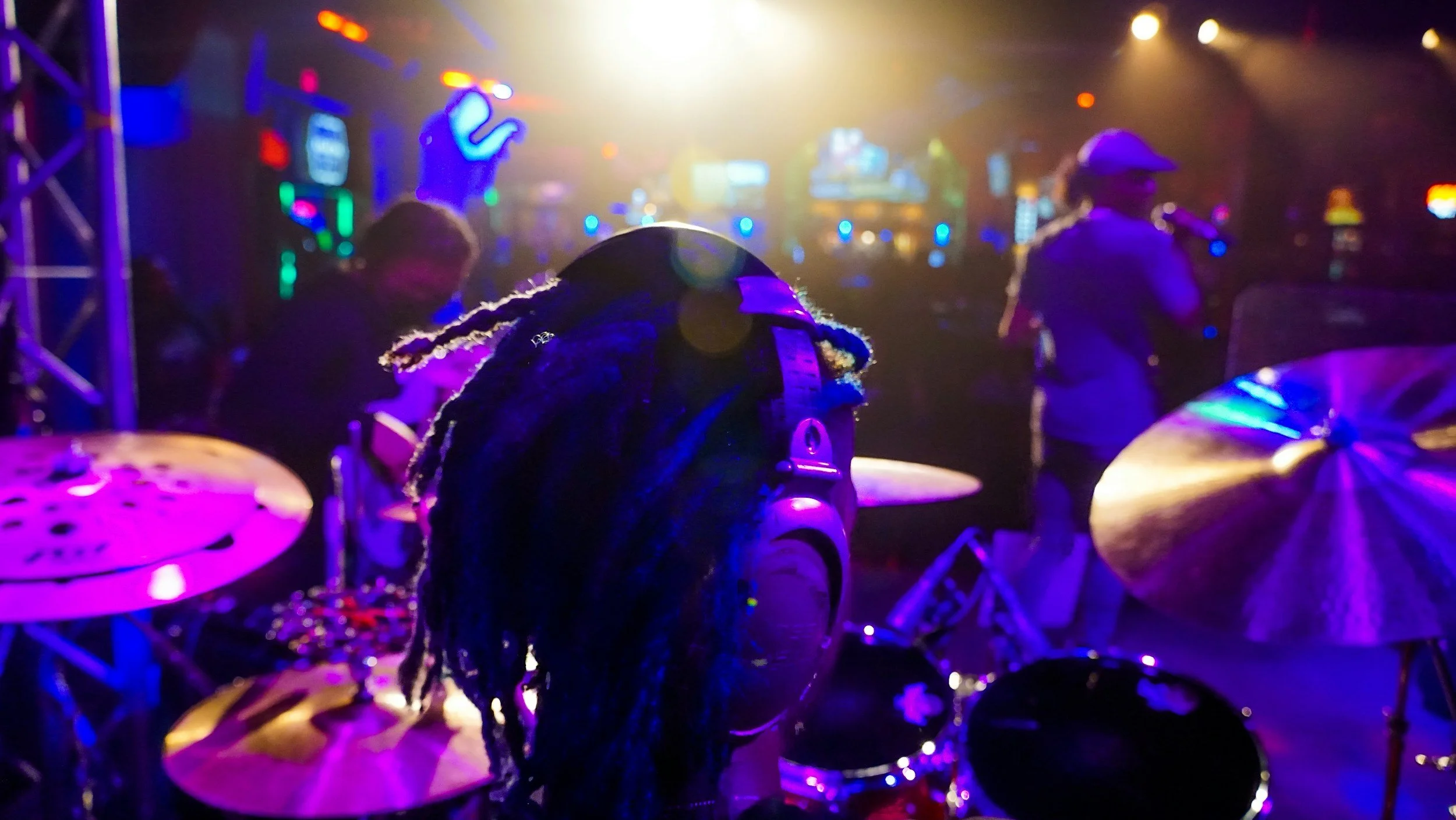 A drummer with dreadlocks is on stage in a dimly lit venue with colorful tonight lights. In the background, there is a female singer and another musician, and various DJ and neon signs.