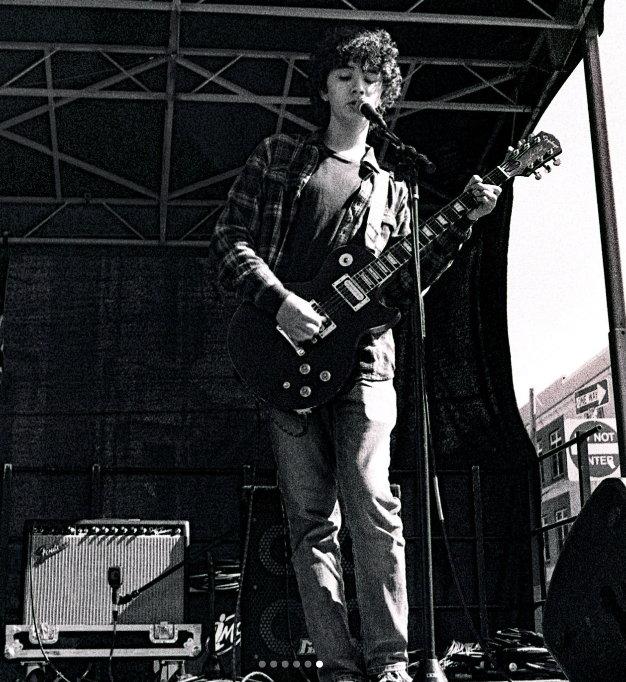 A young person with curly hair playing electric guitar and singing into a microphone on an outdoor stage.