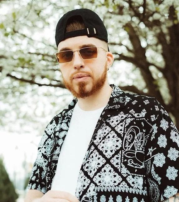 Young man with a beard wearing sunglasses, a backward black cap, a patterned shirt, and a white t-shirt standing outdoors under a tree.