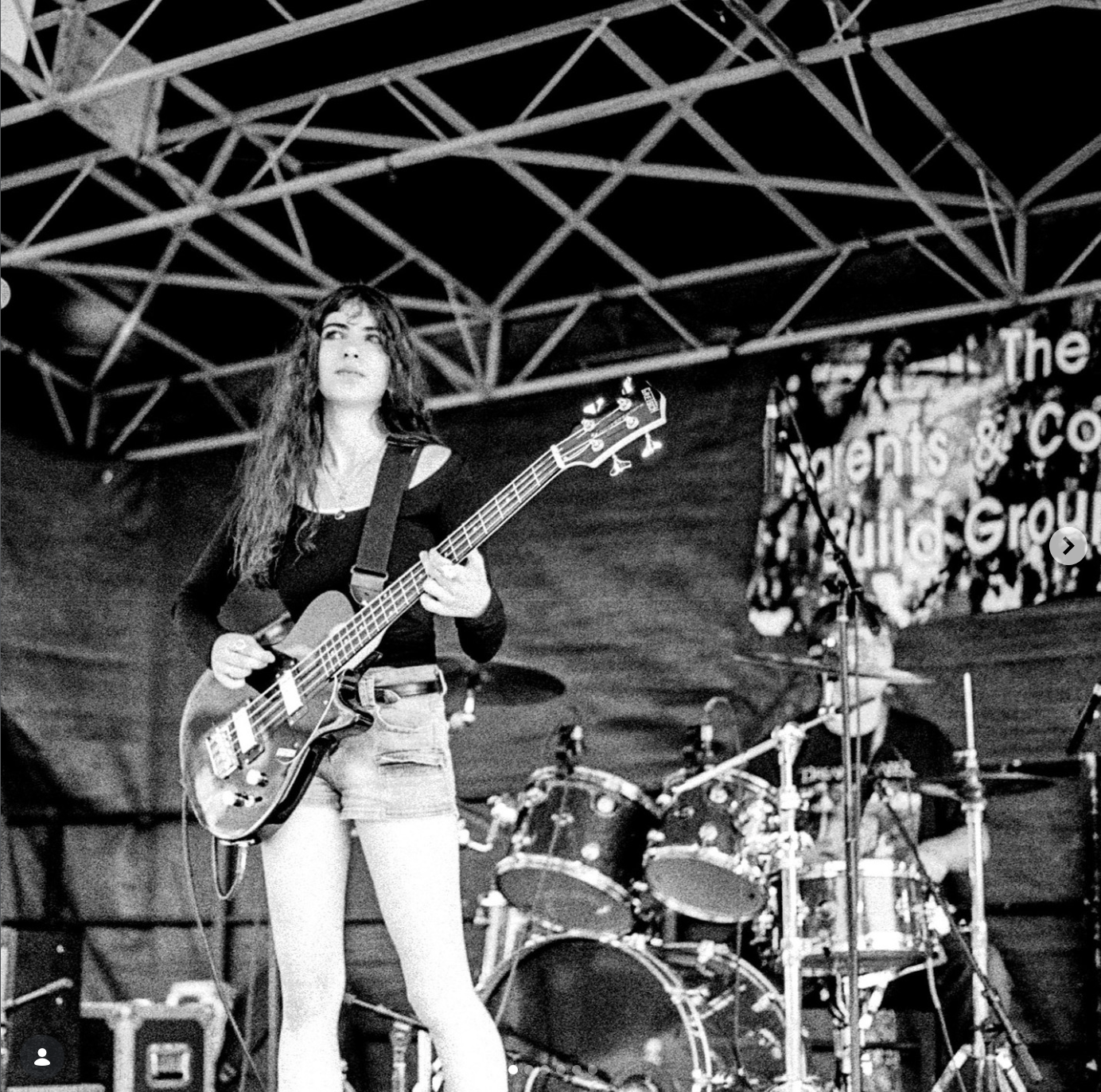 A young woman with long hair plays an electric bass guitar on stage, with a drummer in the background, at a live music event.