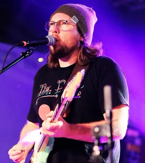 A male musician with long hair, glasses, and a beanie hat singing into a microphone while playing an electric guitar on stage with colorful purple and blue lighting.