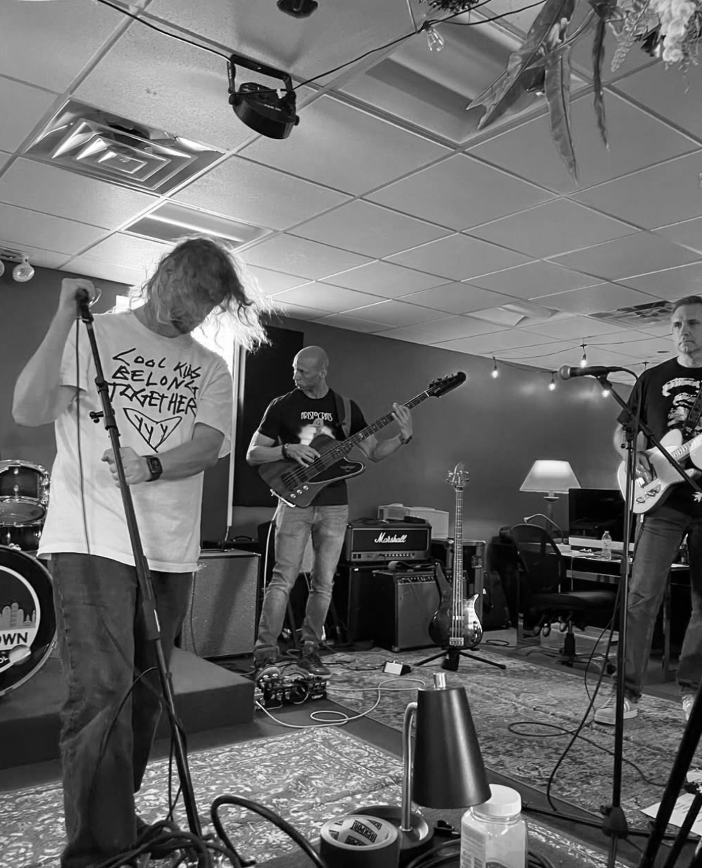 A band practicing in a room with three male musicians playing instruments, one singing into a microphone, a bass guitar, and an electric guitar. There are various equipment and a cabinet in the background.