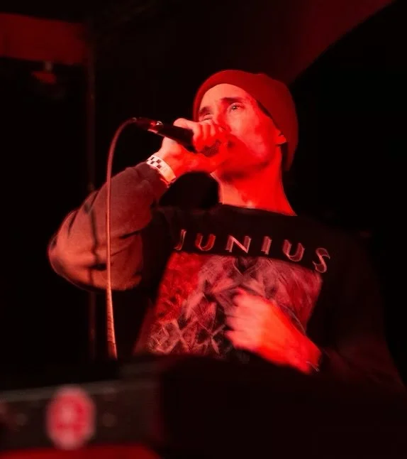 A man wearing a red beanie and a hoodie with a graphic design is singing into a microphone on stage, surrounded by red lighting.
