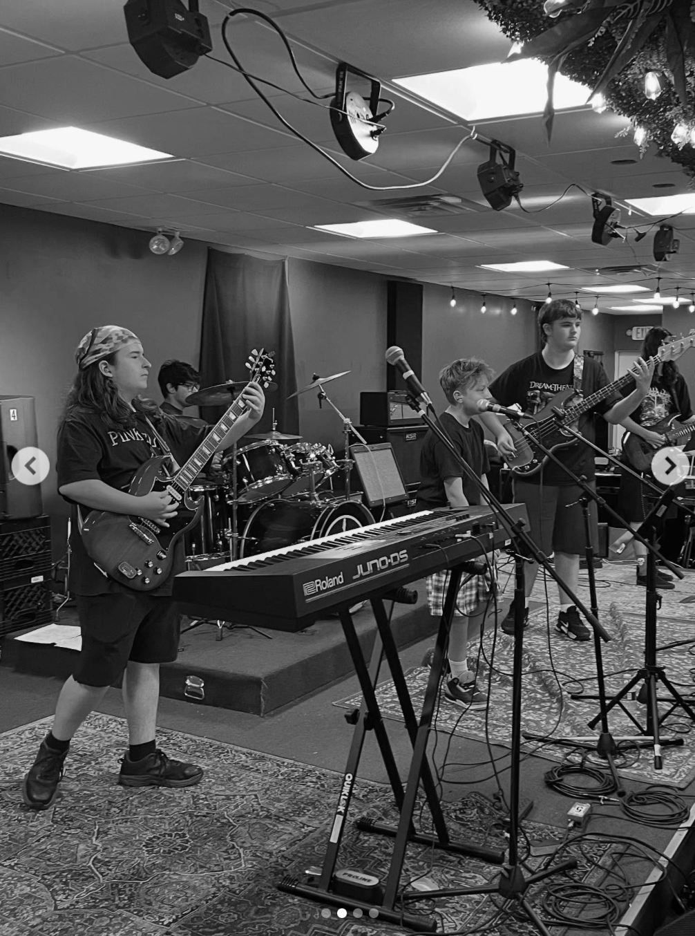 Young children playing musical instruments, including guitar, drums, keyboard, and bass, on stage in a band, during a performance in a decorated indoor setting with string lights and an exit sign.