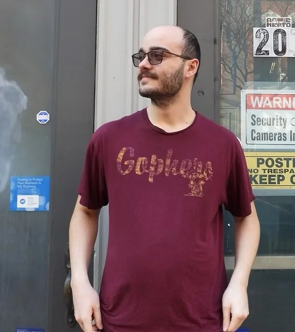 Man with glasses and beard wearing a maroon Gadsden T-shirt standing outside near a door with security and warning signs.