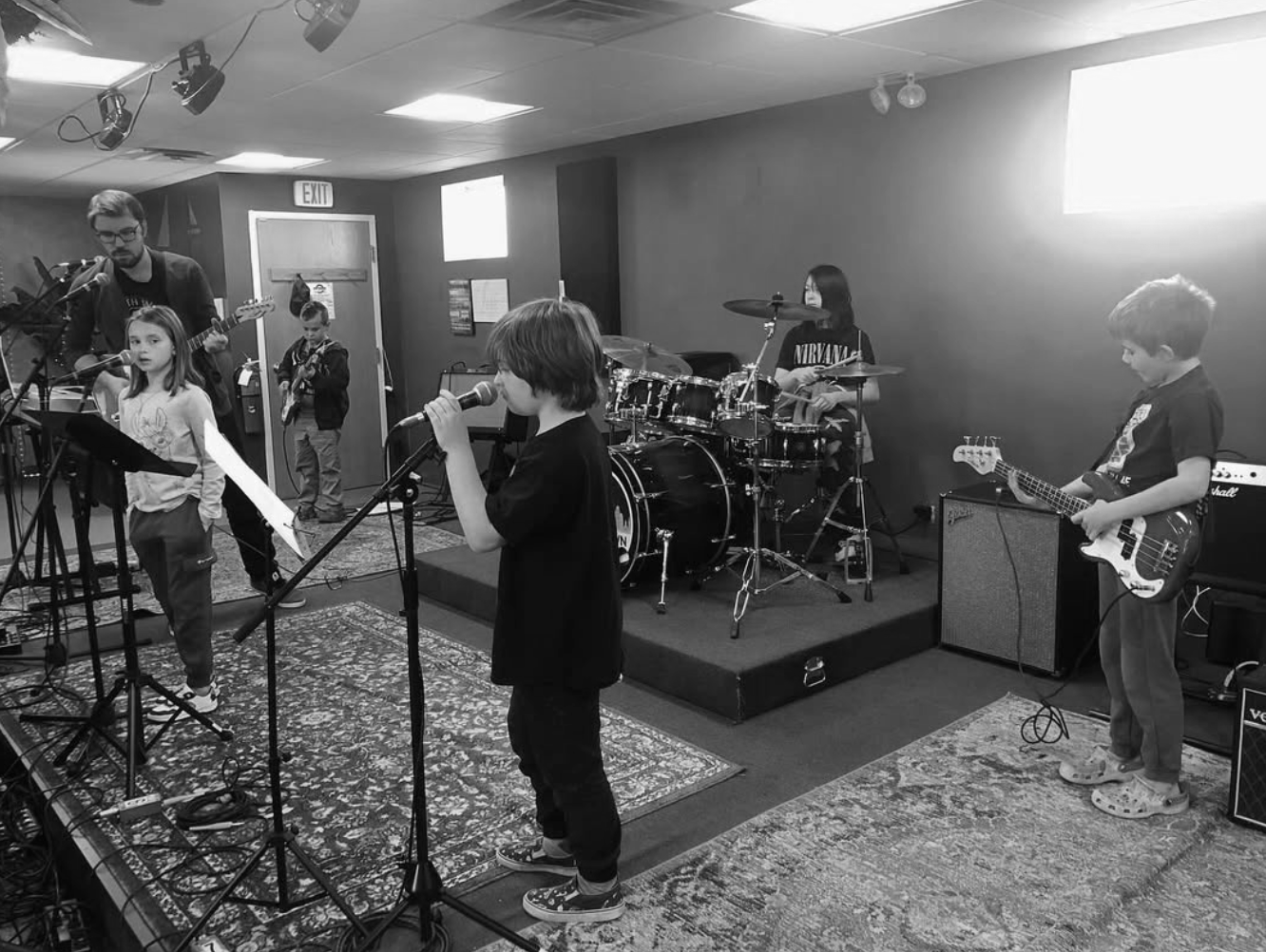 Children performing in a band with microphones, guitars, and drums in a room.