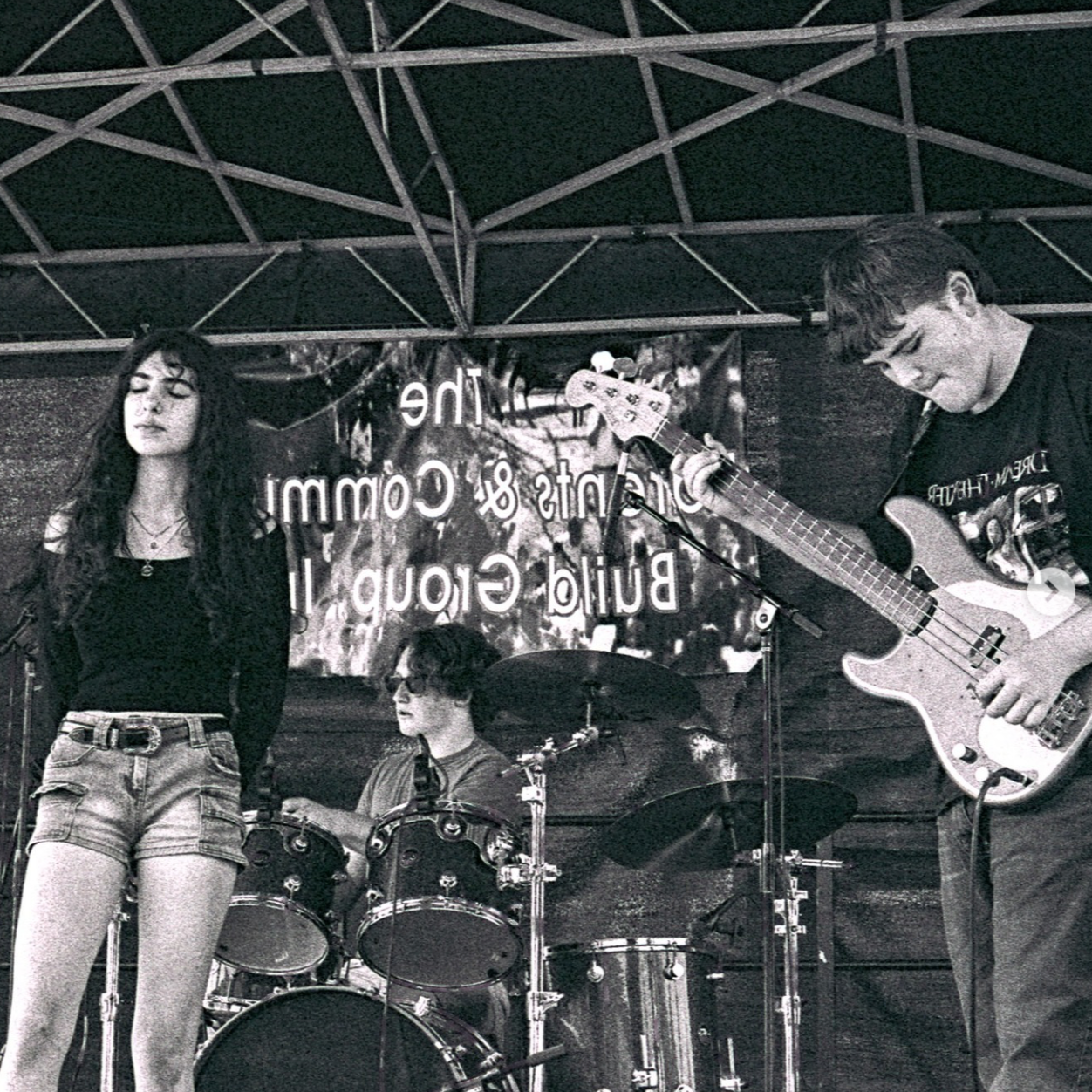 A black and white photo of three young musicians performing on an outdoor stage. The female singer is standing on the left with her eyes closed, wearing a black top and denim shorts. The male guitarist on the right is playing an electric guitar, wearing glasses and a dark T-shirt. The drummer in the background is sitting at a drum kit with sunglasses.