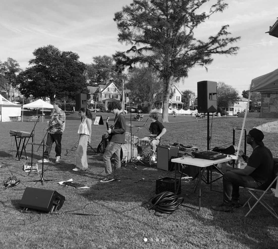 A group of musicians performing outdoors on a grassy field, with trees and houses in the background, and tents set up nearby.