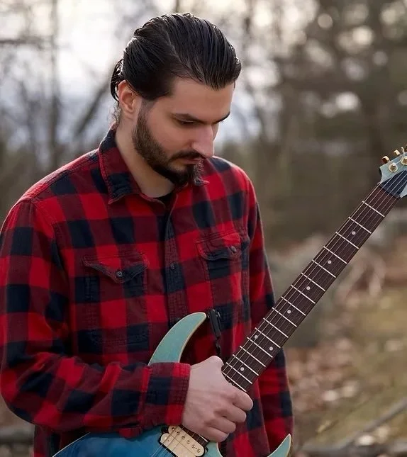 A man with dark hair and a beard holding an electric guitar outdoors, wearing a red and black checkered flannel shirt.