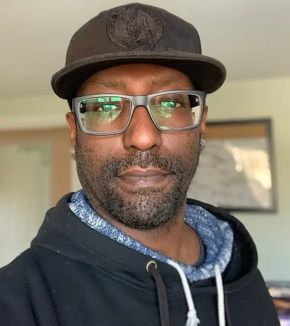 A man with glasses and a black baseball cap. He is wearing a hoodie and has a beard and mustache.