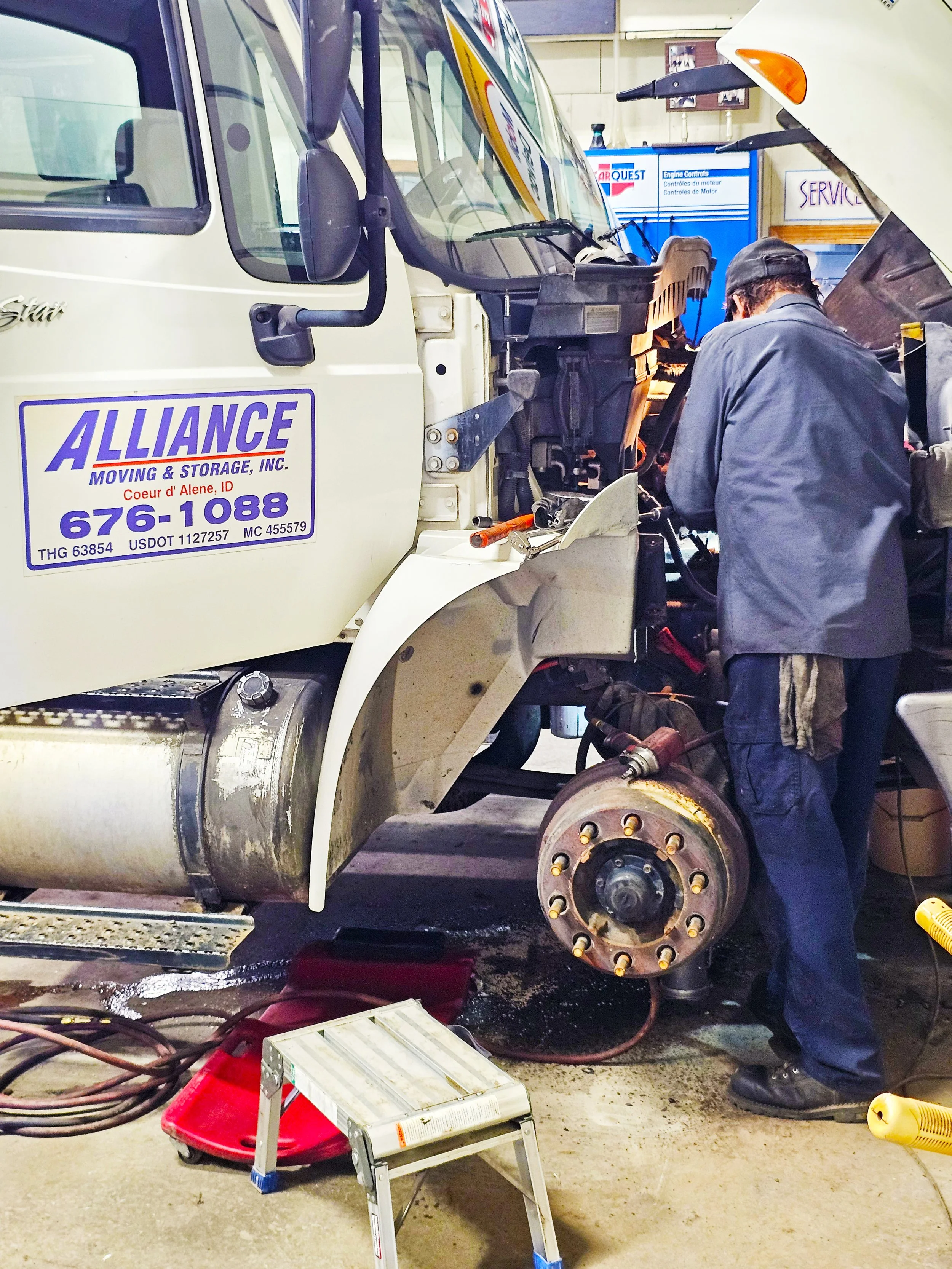 A mechanic working on a large truck in an auto repair shop, with the front wheel removed and tools around.