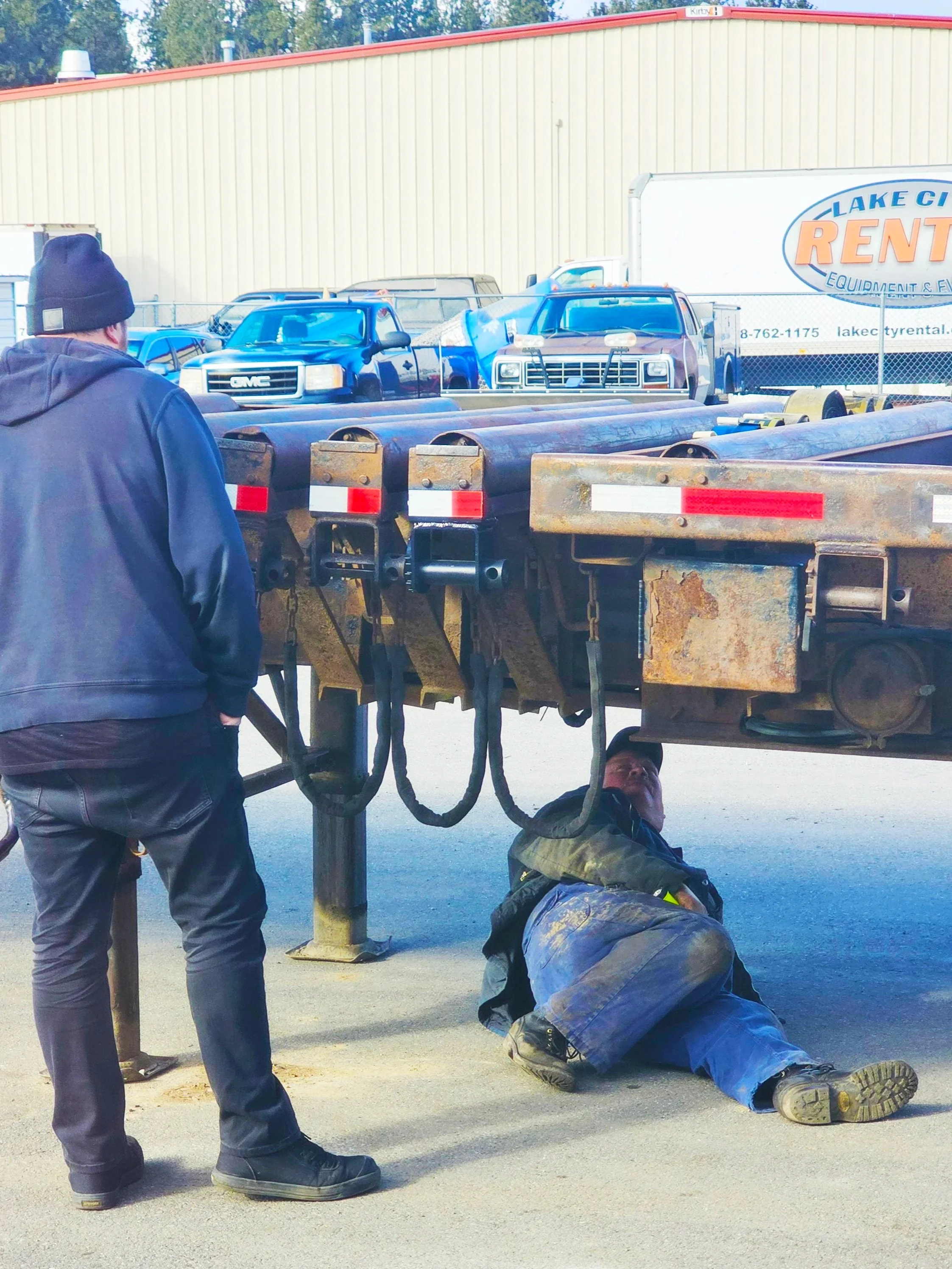 A person is working under a flatbed trailer, while another person oversees the work. The scene is outdoors in a parking lot with other trucks, RVs, and trailers with a building in the background.