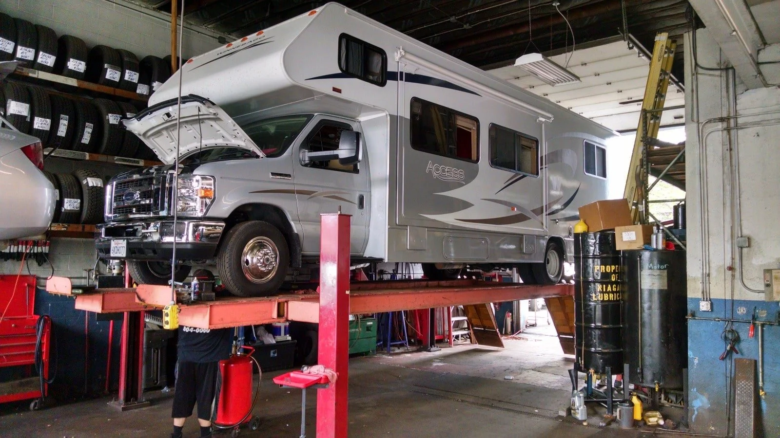 A recreational vehicle (RV) is lifted on a hydraulic repair lift inside a garage or workshop. The RV's hood is open, indicating maintenance work.
