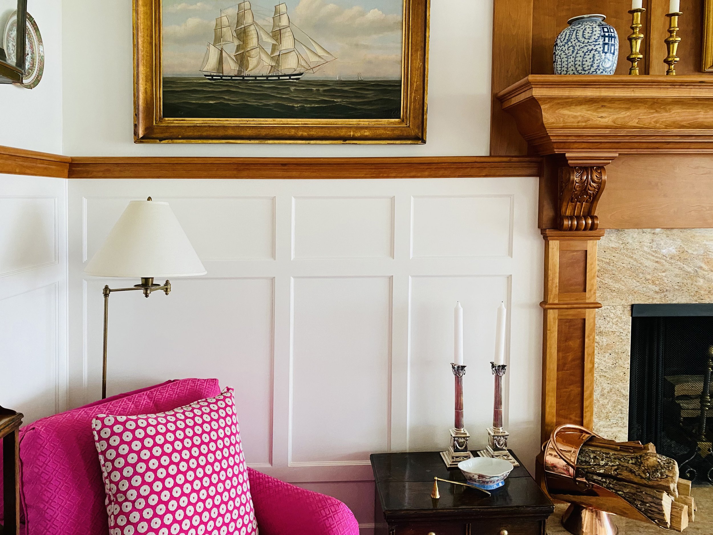 Living room with a white paneled wall, wooden mantel, a painting of a ship, a pink armchair with a patterned pillow, a gold lamp, a black side table with two candlesticks, and a fireplace with a wood log holder.