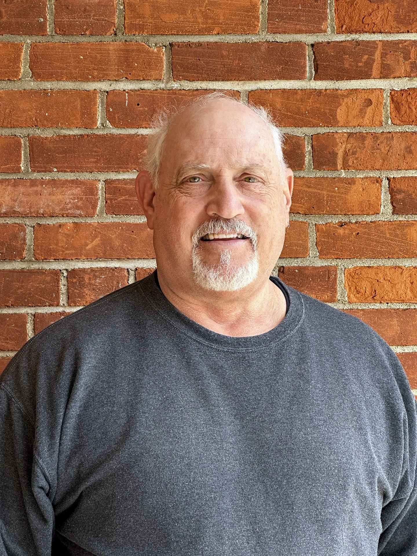 A man with a white beard and mustache, smiling, wearing a dark gray sweatshirt, standing in front of a red brick wall.
