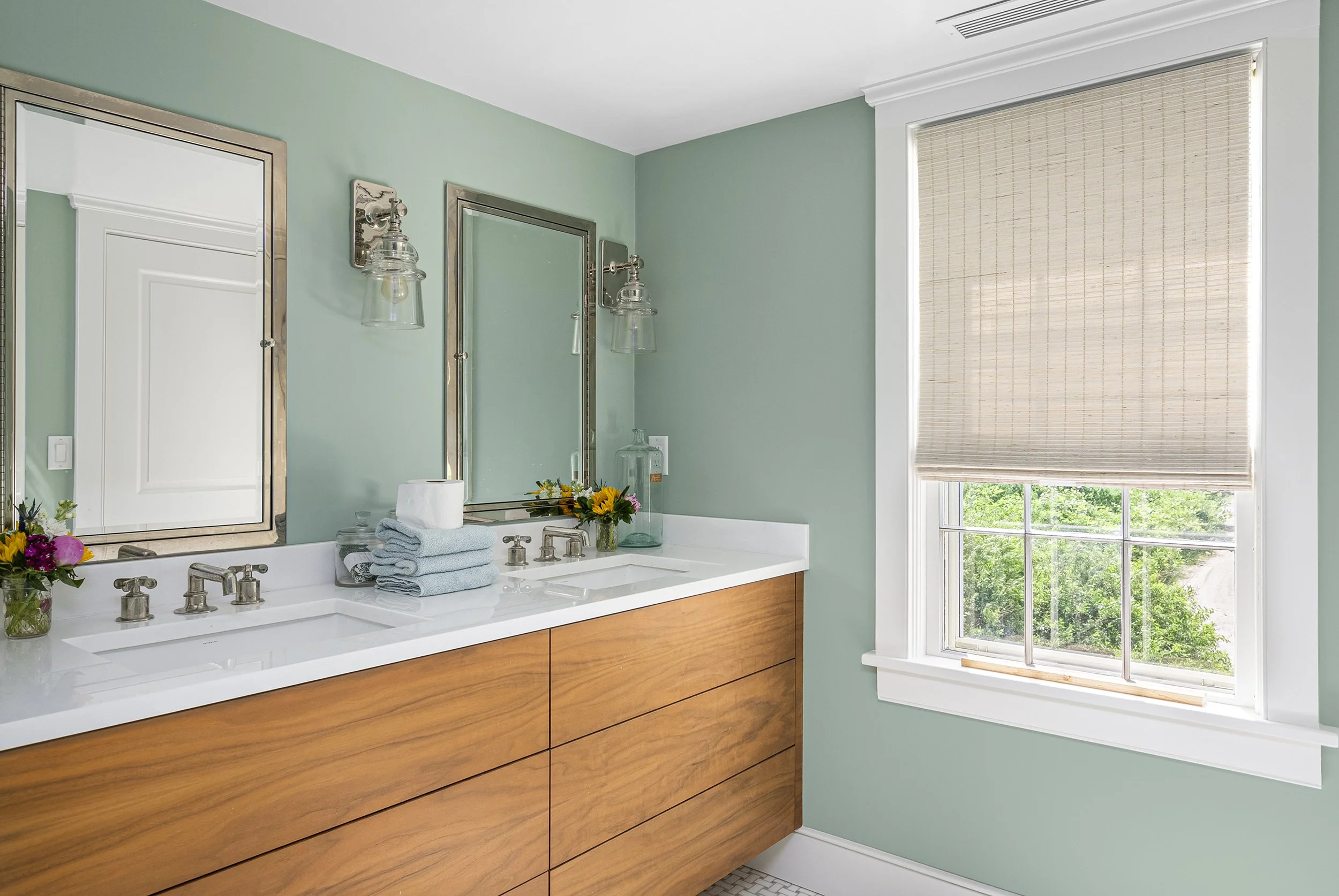 Bathroom with green walls, double white sink vanity with wood drawers, two large mirrors, wall-mounted light fixtures, and a window with a beige shade showing green trees outside.
