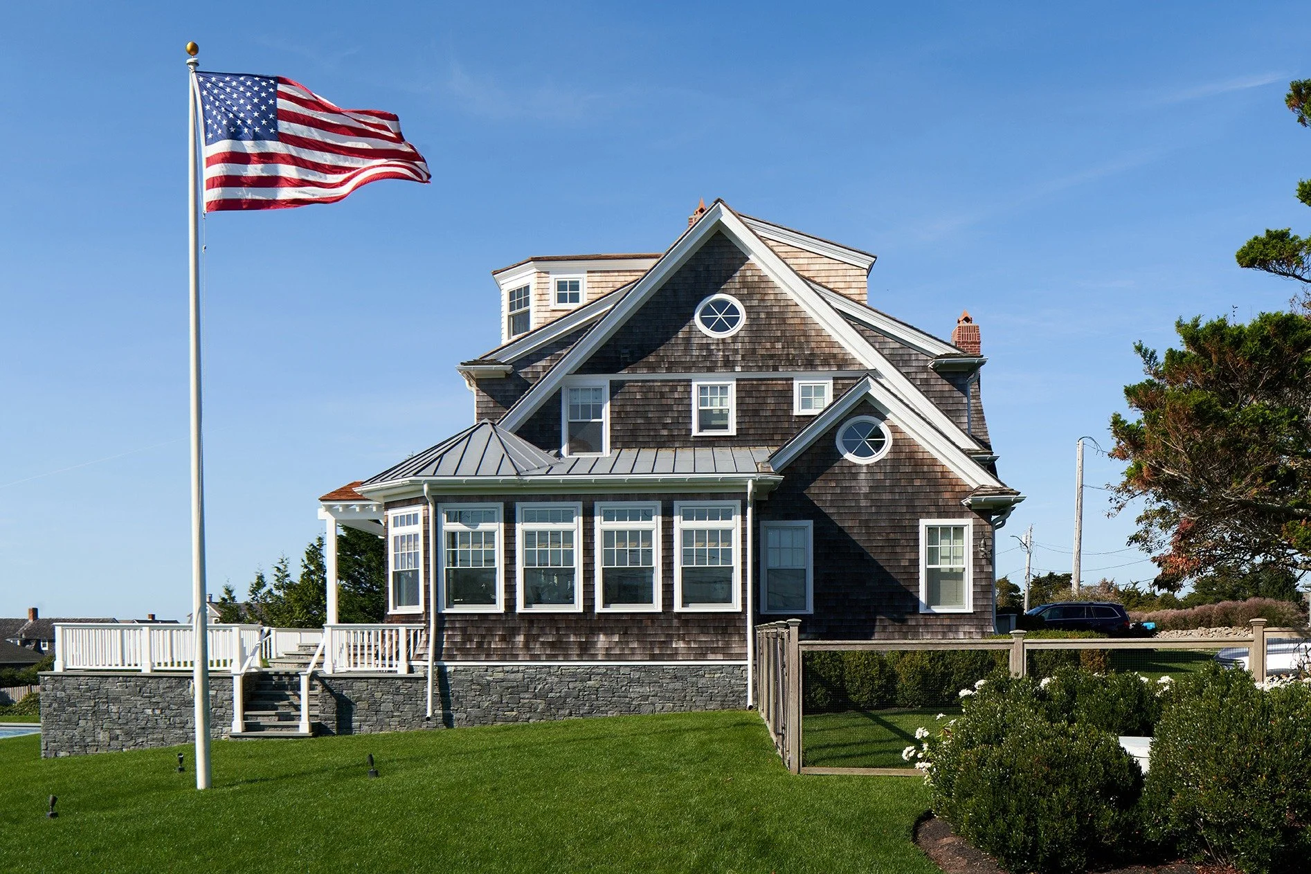 A large, multi-story house with a peaked roof, shingles, and multiple windows. An American flag on a flagpole is in the foreground, and a well-manicured lawn surrounds the house. There are a few trees and a clear blue sky in the background.