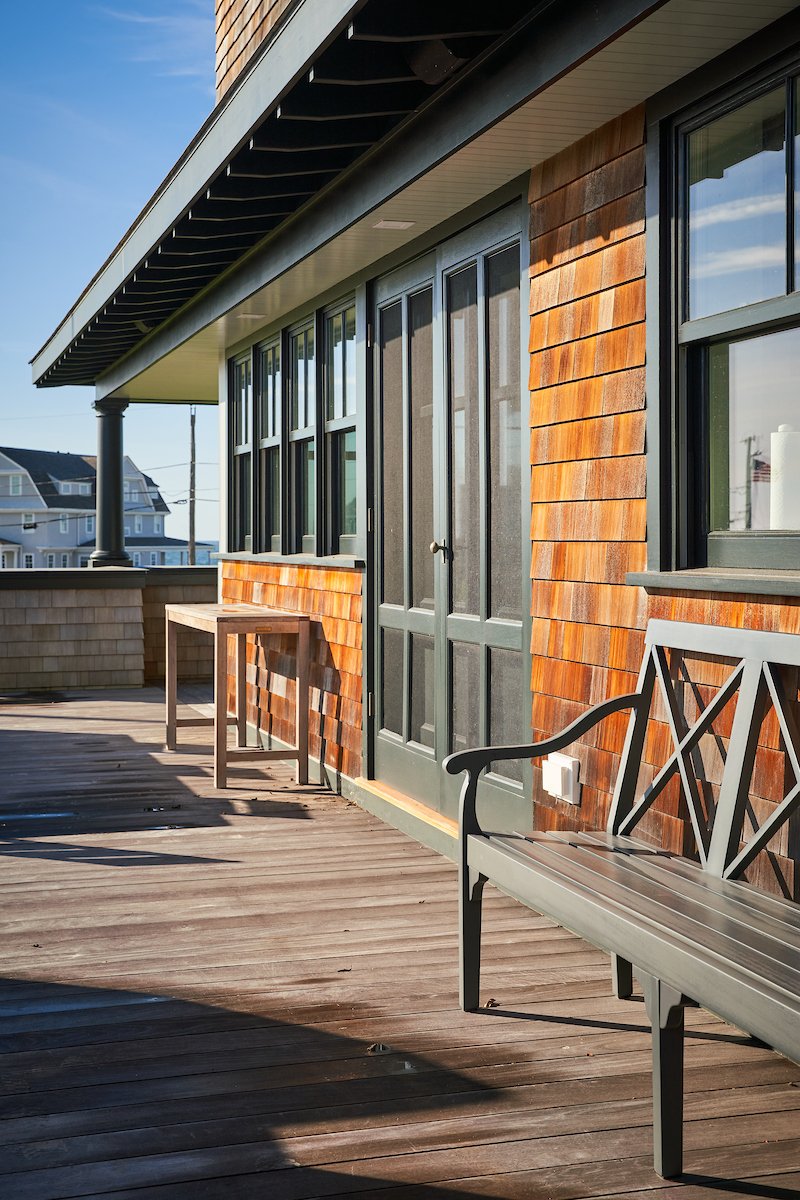 A wooden balcony with a gray bench, small table, and glass-paneled doors attached to a house with a brown shingle exterior. The sky is clear and blue, and neighboring houses are visible in the background.