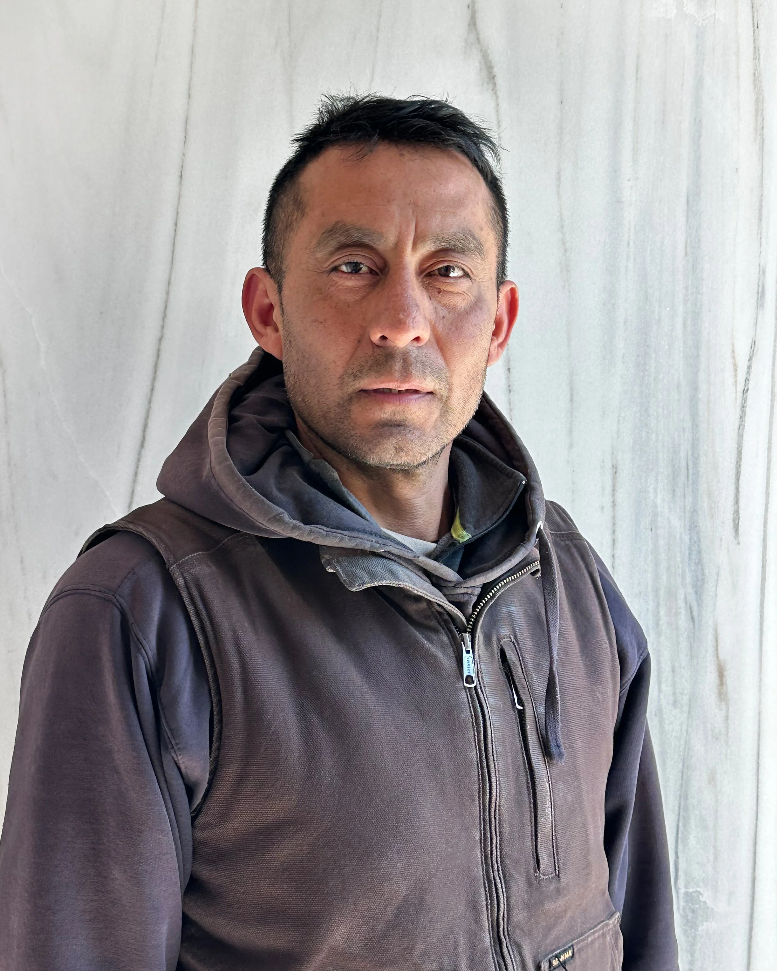 Close-up of a man with short dark hair, wearing a brown zip-up jacket with a hood, standing in front of a plain, light-colored wall.