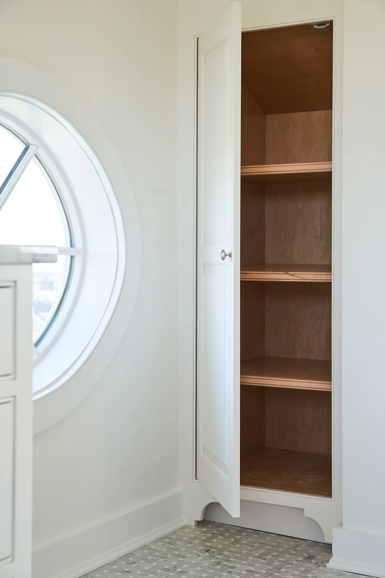Empty wooden closet with white frame next to a round window in a room with white walls and mosaic tile floor.