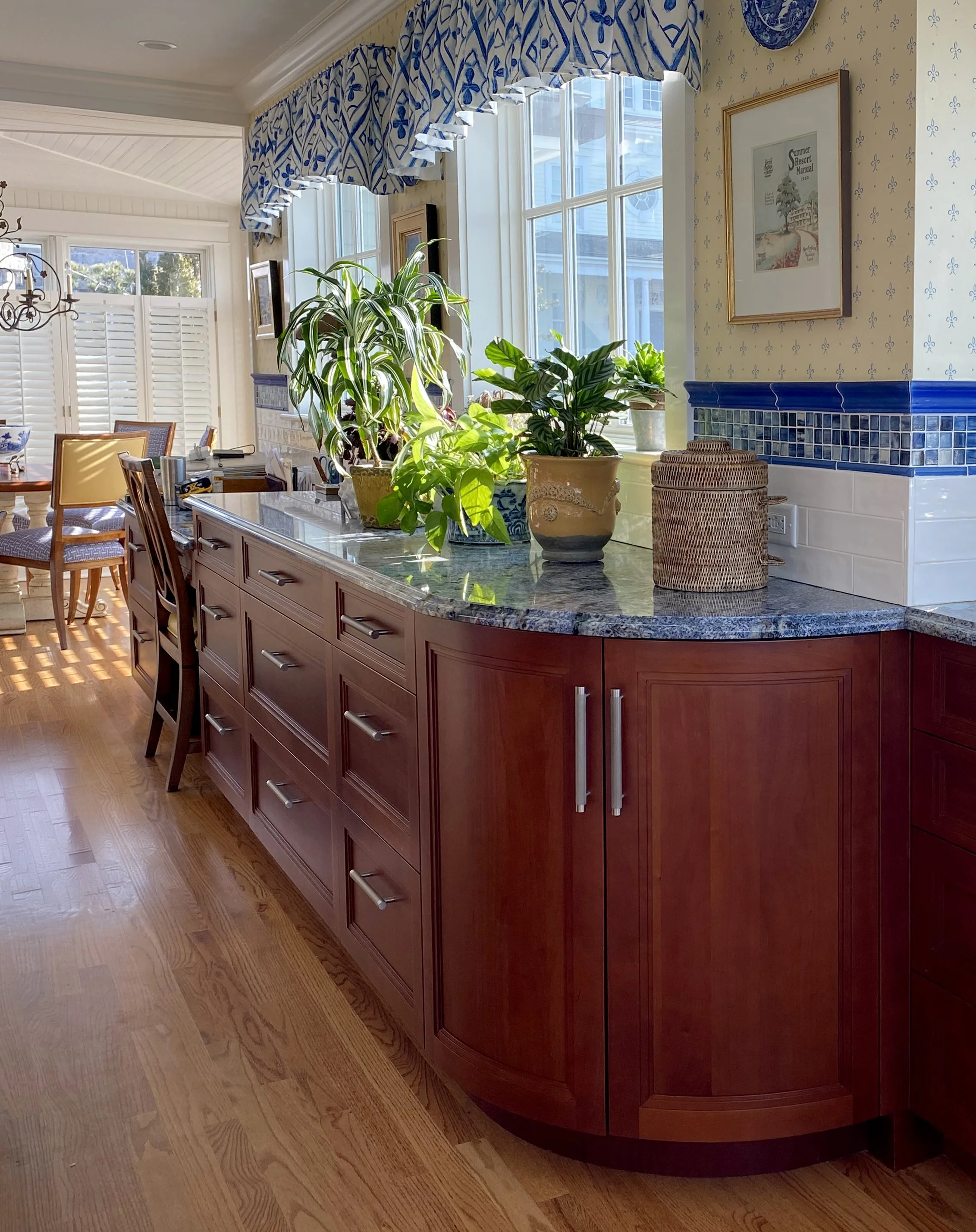 A kitchen with wooden cabinets, a granite countertop, potted plants near a window, and a dining area in the background.