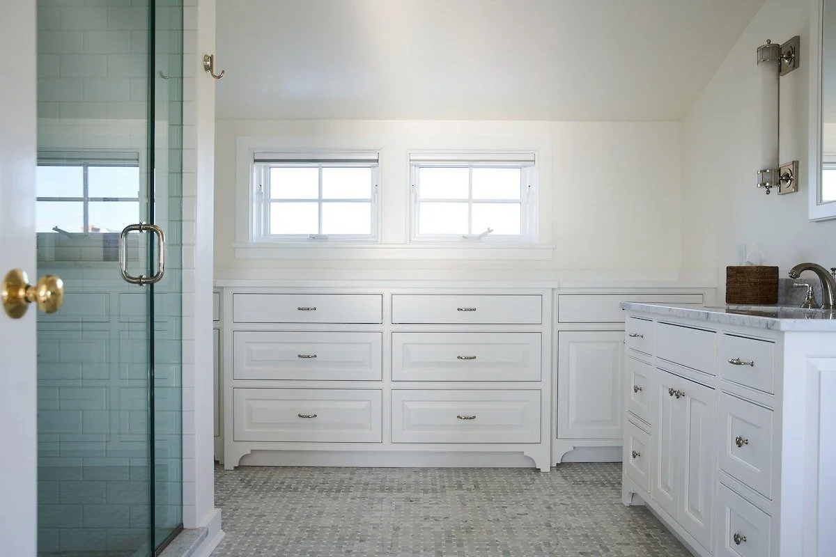 Bright bathroom with white cabinets, two small windows, and a glass shower door on the left.