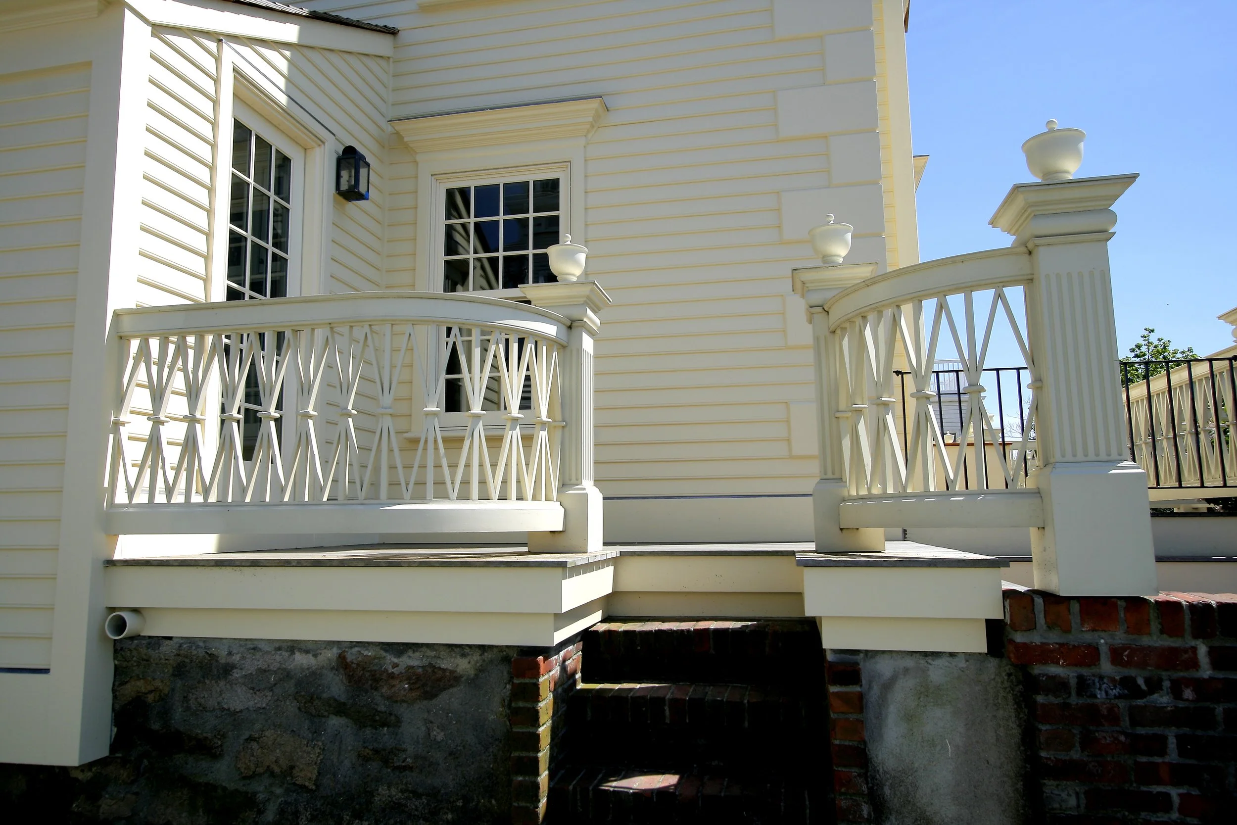 A cream-colored house with a small elevated porch featuring decorative white railings and columns, set against a clear blue sky.