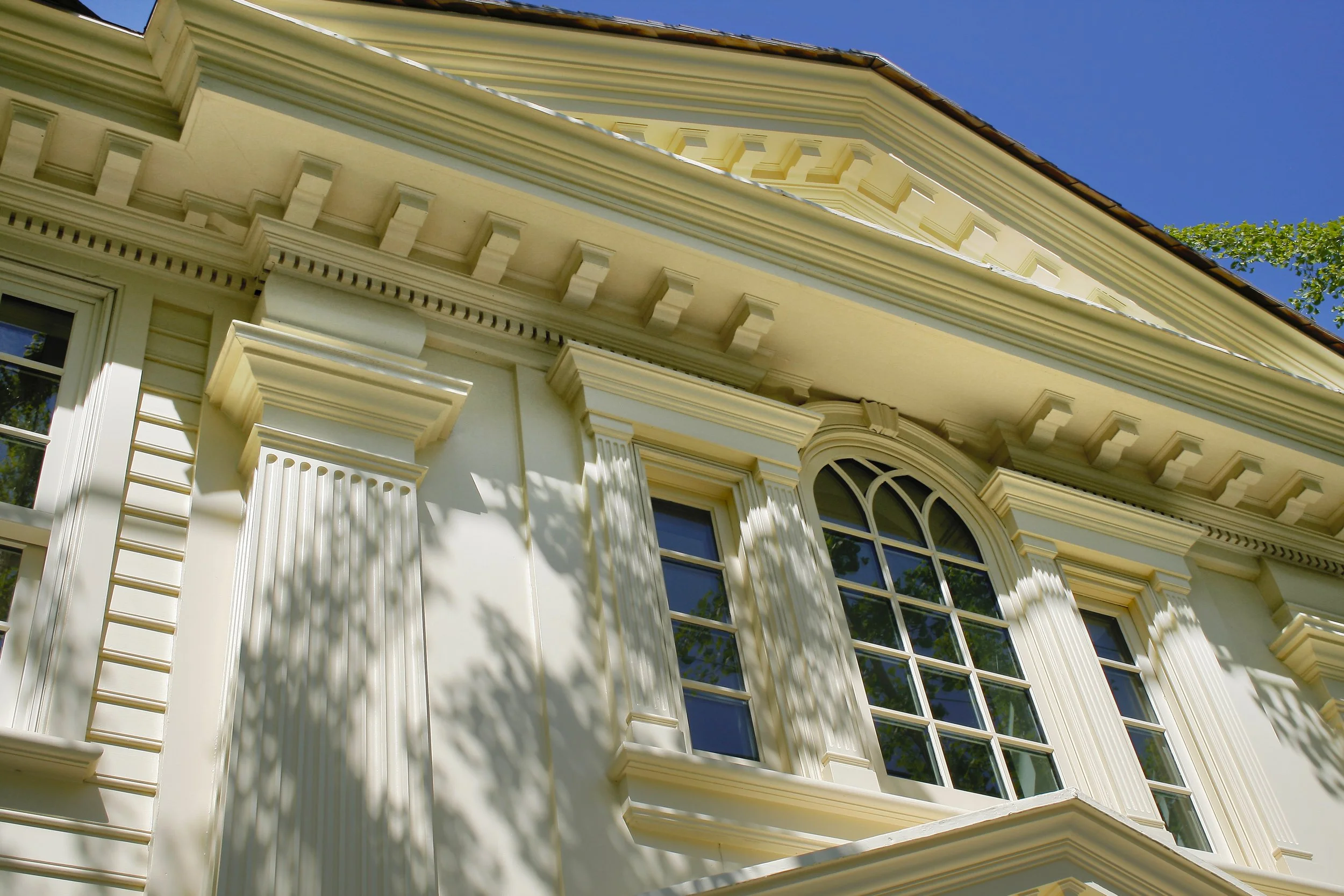 Close-up of a cream-colored house exterior with decorative moldings, large arched window, and attic vent, under a clear blue sky.