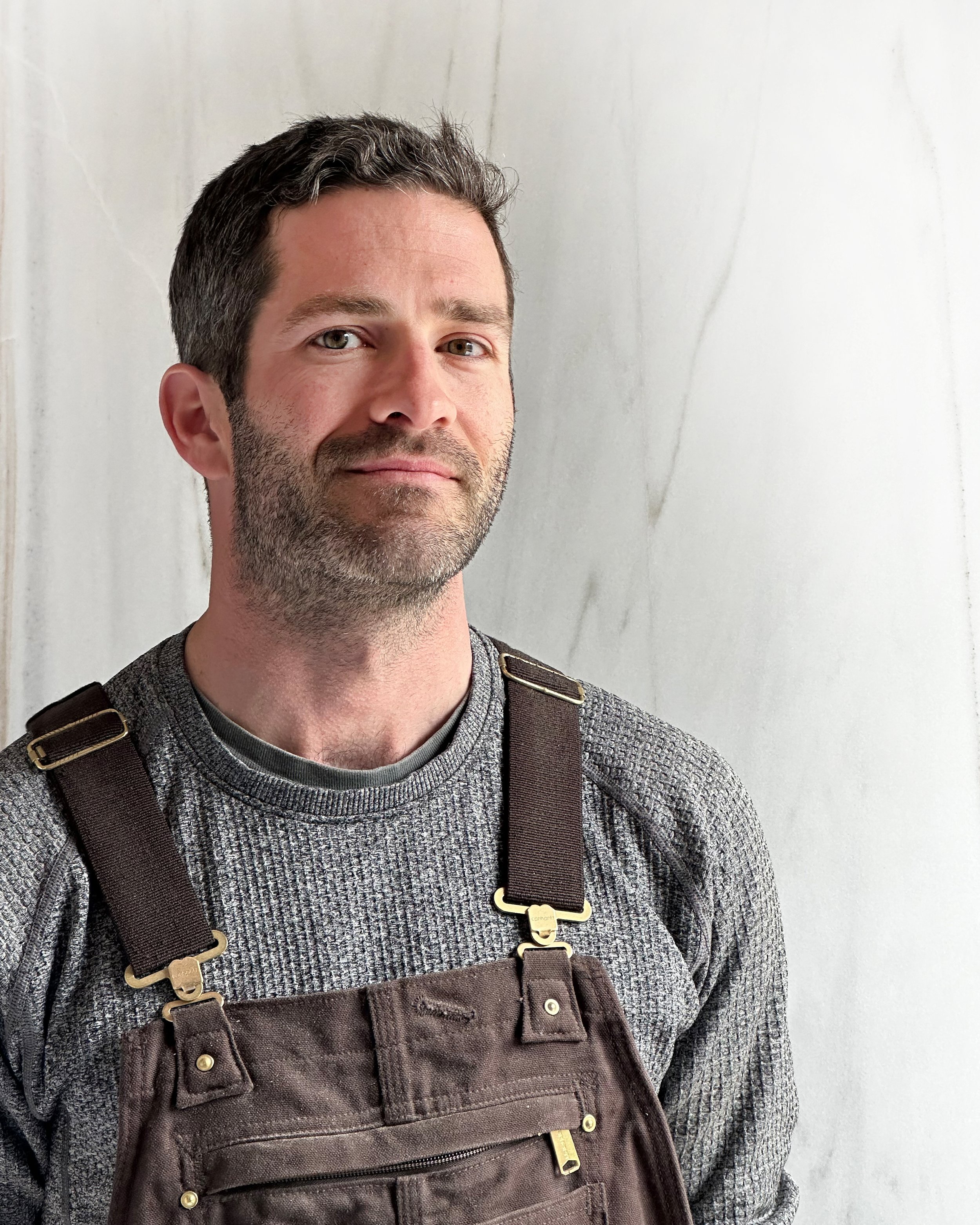 A man with dark hair, facial hair, and blue eyes, wearing a gray sweater and brown overalls, stands against a white wall with subtle marble veining.