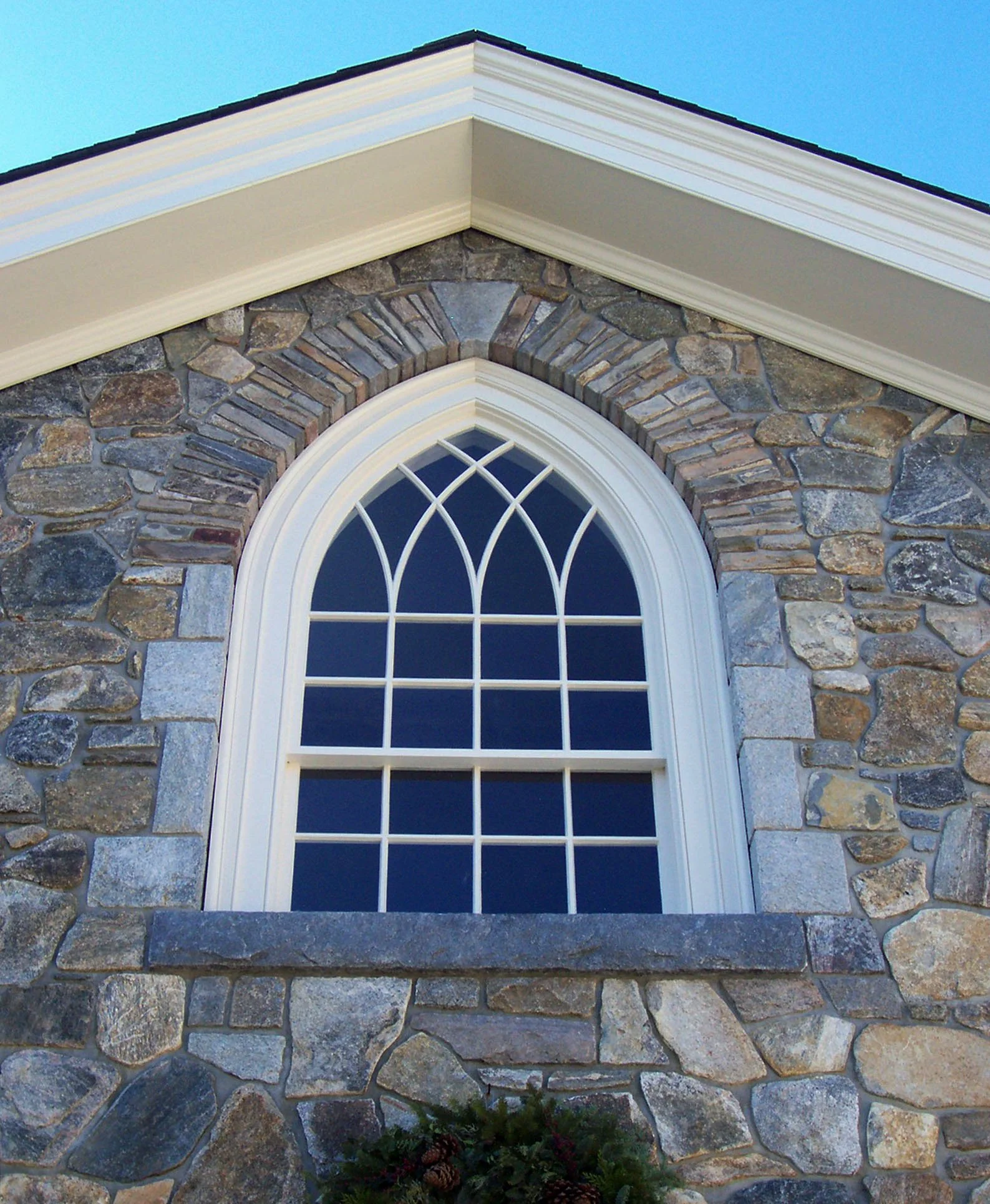 Close-up of a stone house exterior with a large arched window, white trim, and a black shingled roof against a blue sky.