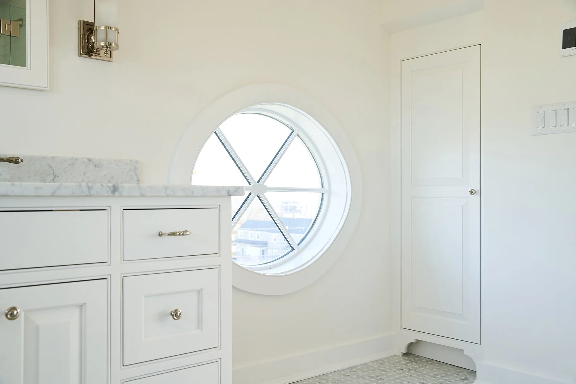 A bright, white bathroom with a circular window, white cabinetry, marble countertop, and a door on the right side.