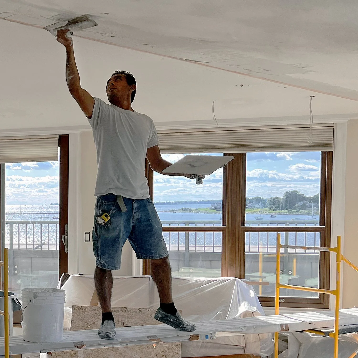 A man in a white t-shirt and shorts stands on a scaffolding, painting the ceiling of a room during home renovation, with large windows showing a view of water and sky outside.