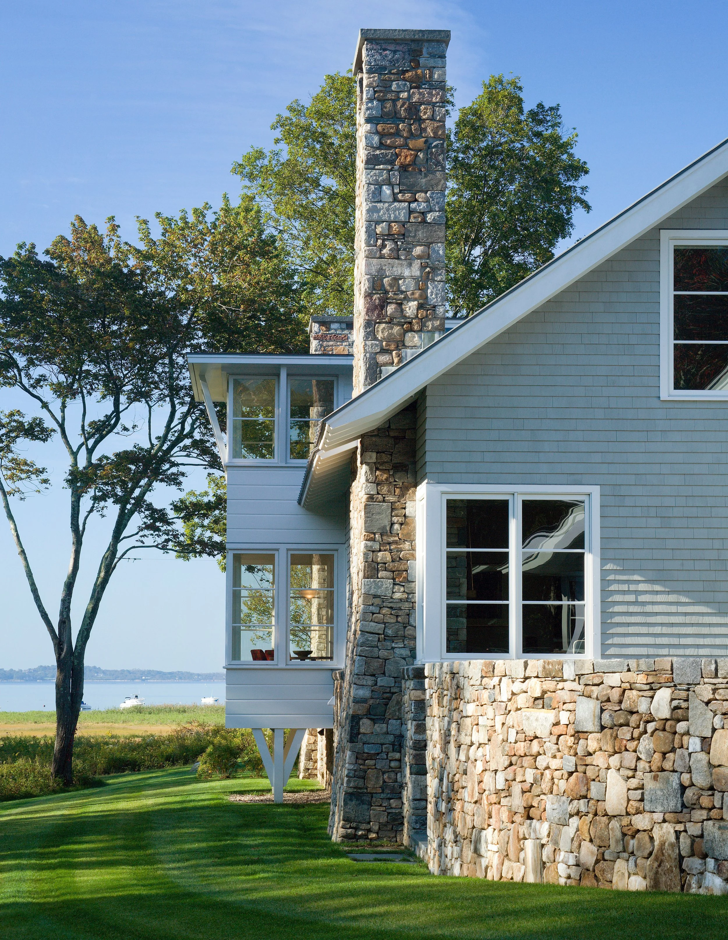A house with a stone chimney, gray siding, and large windows, with a green lawn and trees in the background, near a body of water.