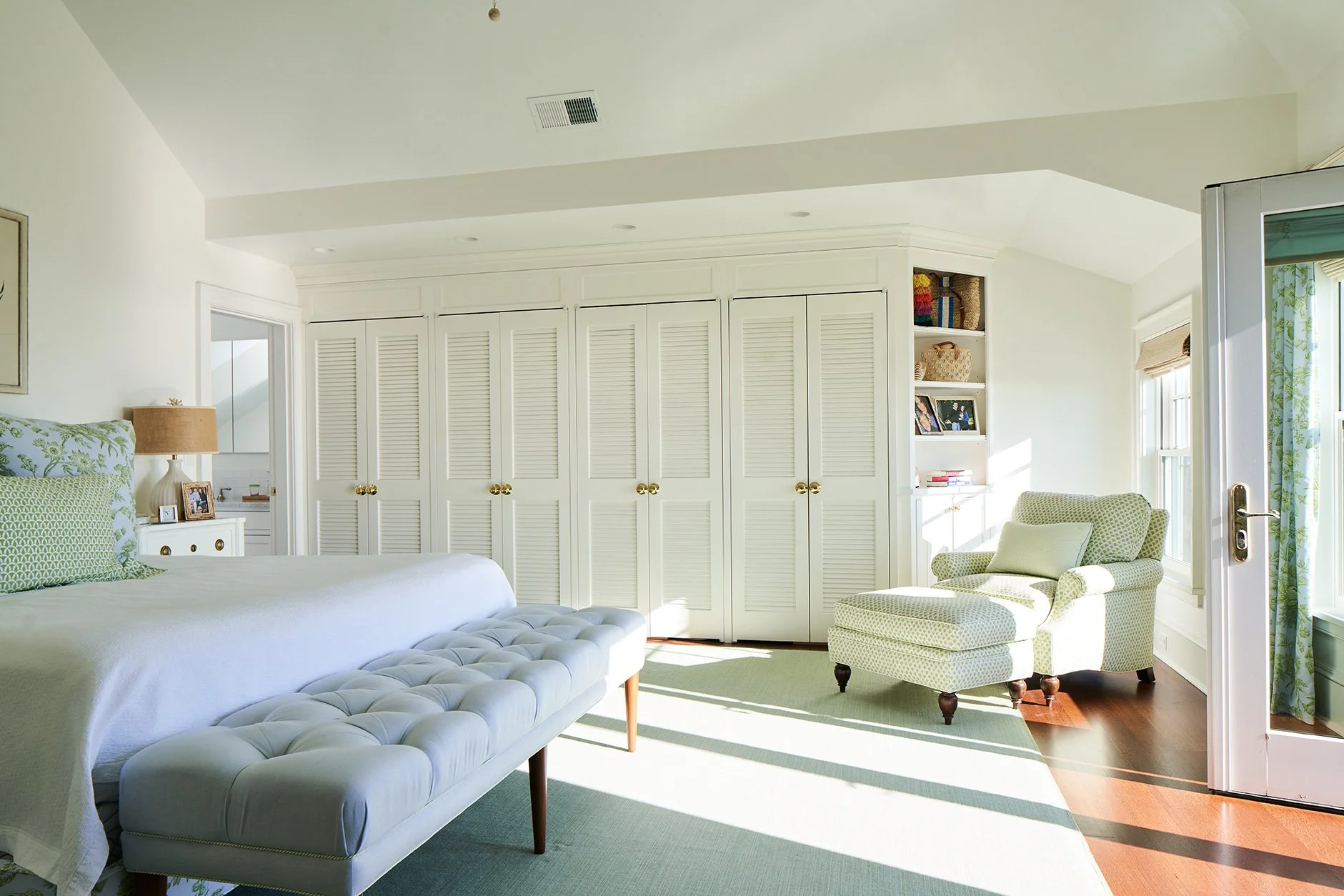 Bright bedroom with white walls, green accents, a large bed with patterned pillows, built-in closet with louvered doors, a white armchair with a footrest near a window, and a door to outside with floral curtains.