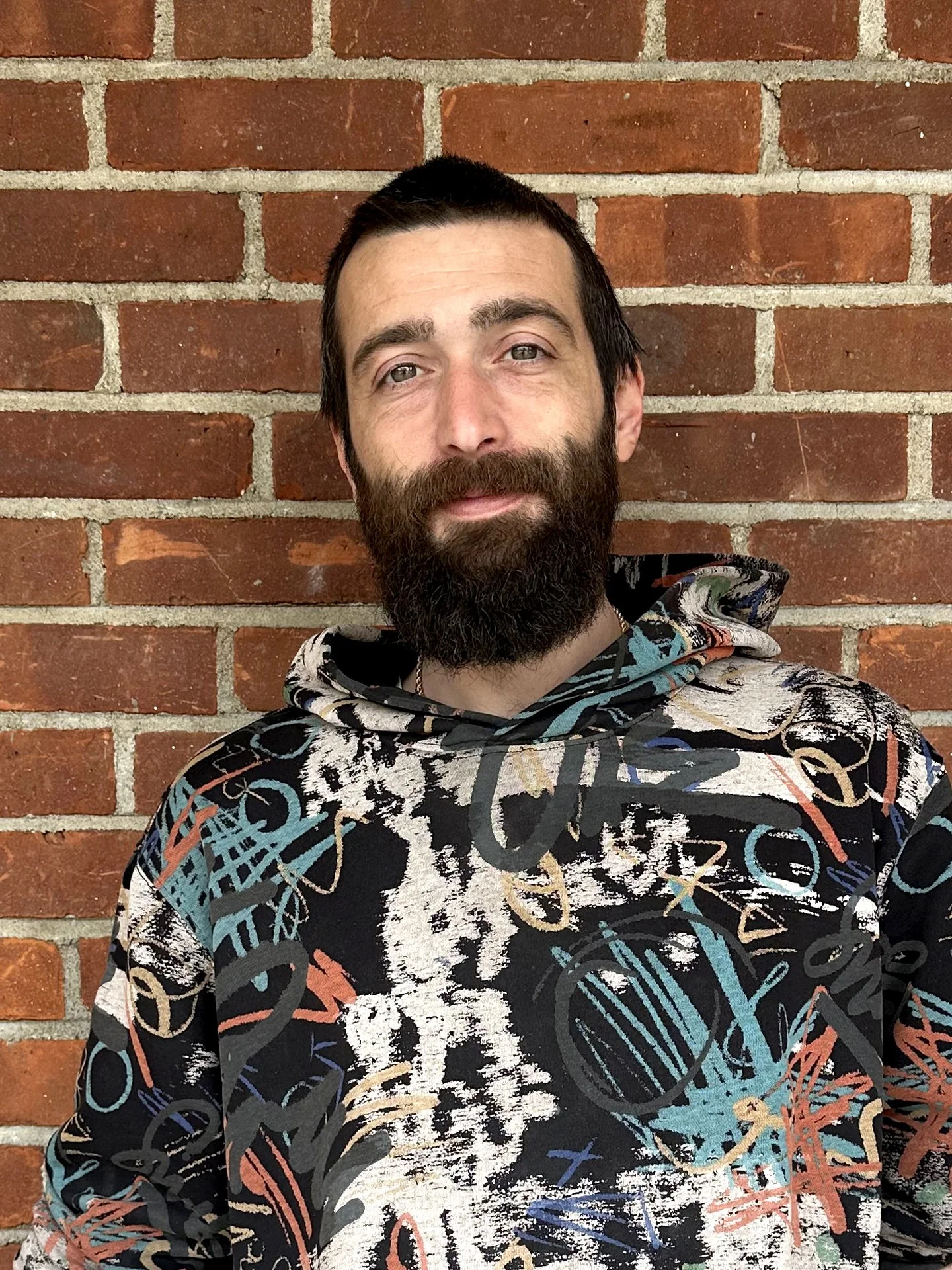 A man with a dark beard and short dark hair standing in front of a red brick wall, wearing a colorful abstract patterned hoodie.