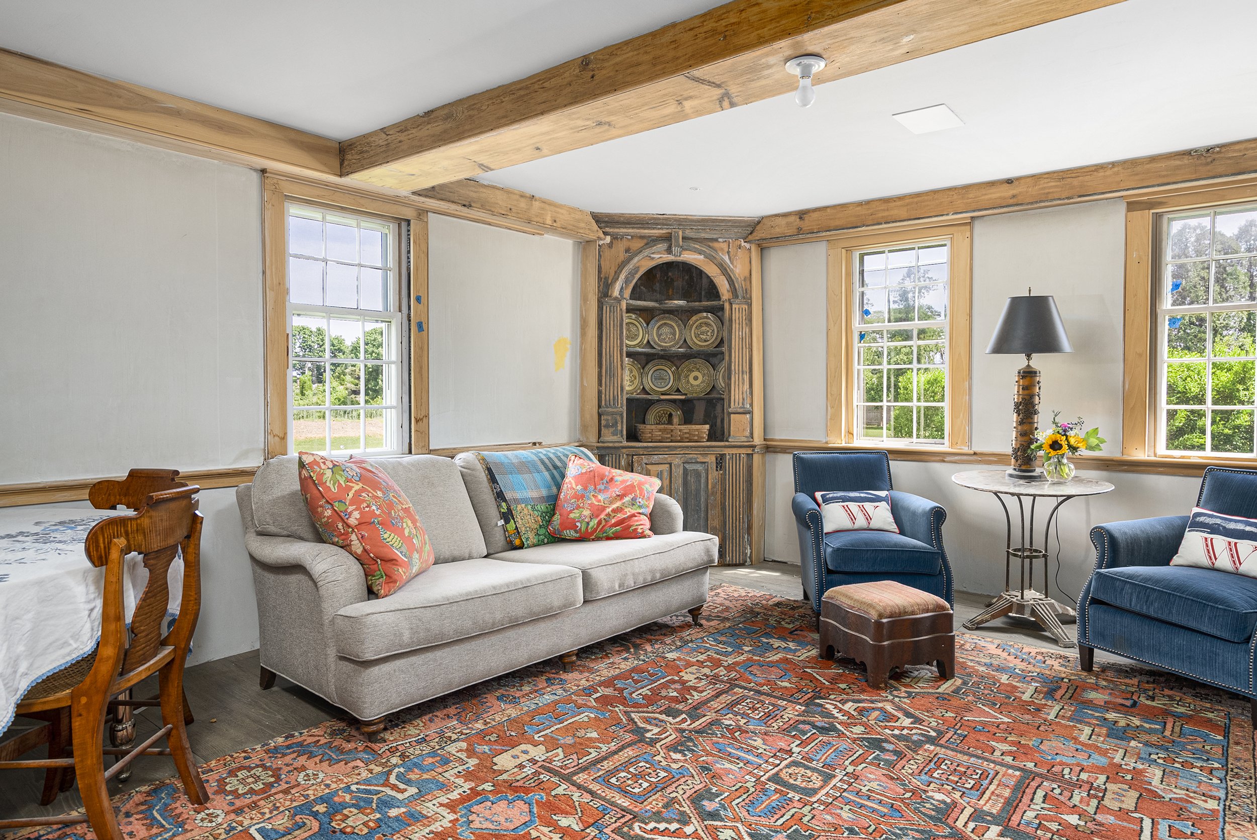 Living room with wooden trim, beige walls, and three windows showing trees outside. Contains a gray sofa with colorful pillows, two blue armchairs with pillows, a small round table with a lamp and sunflower arrangement, and an ornate cabinet in the c