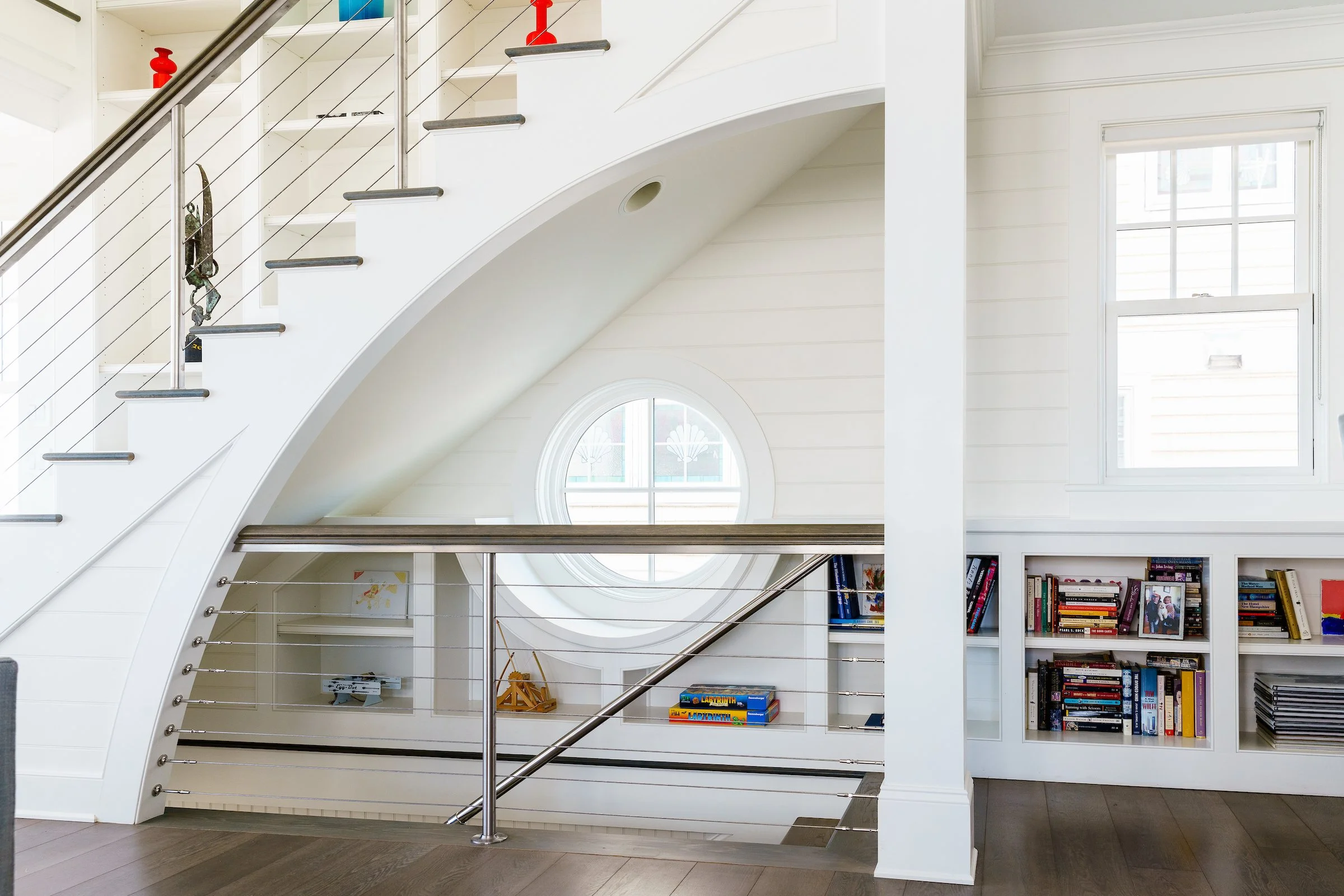 Interior of a modern house with white walls, a staircase with metal railing, large windows, and a built-in bookshelf filled with books.