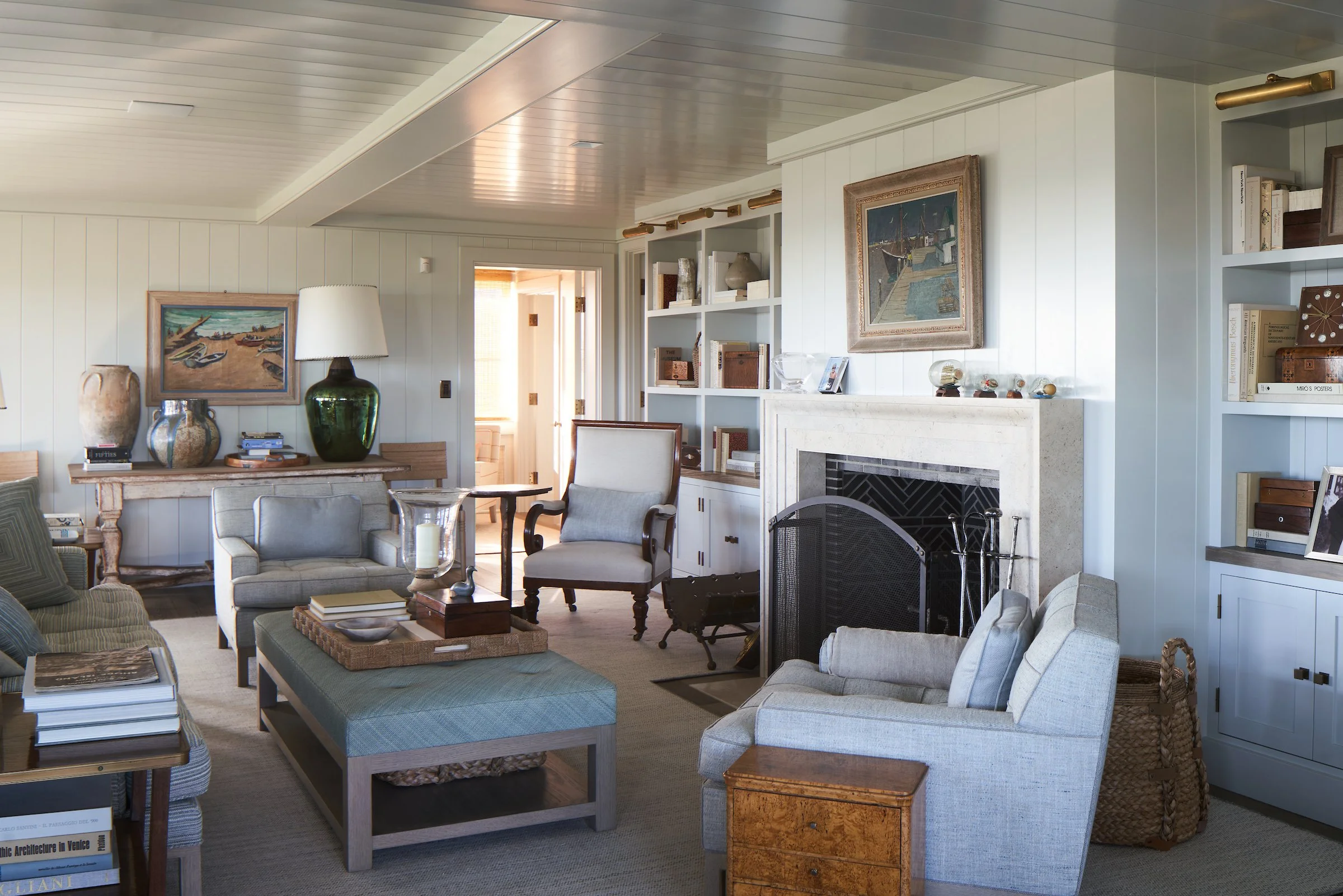 A cozy living room with light-colored walls and ceiling, featuring a fireplace with a white mantel, built-in bookshelves filled with books and decorative items, and comfortable seating including armchairs and a sofa. There are paintings and vases on 