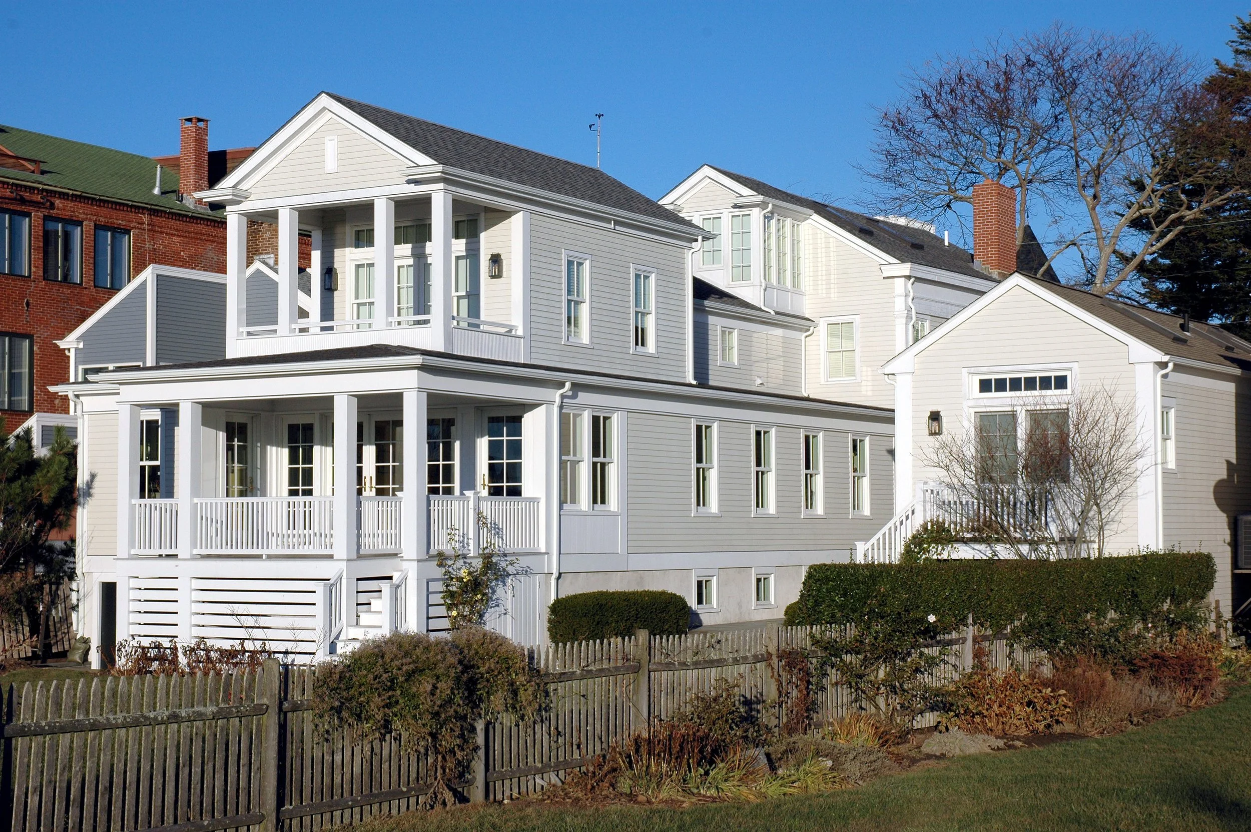White multi-story house with a porch and multiple windows, surrounded by a wooden fence, bushes, and trees, under a clear blue sky.