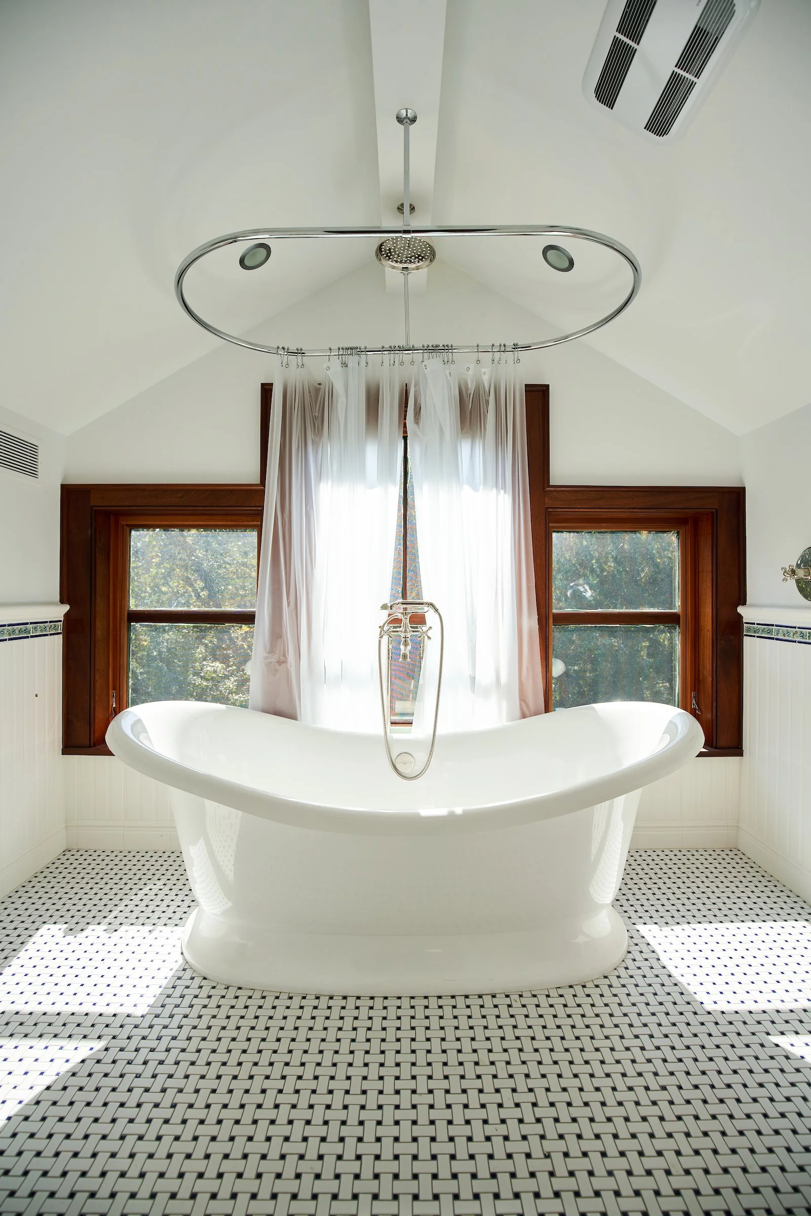 Bright bathroom featuring a freestanding bathtub, two windows with wooden frames, white curtains, and a ceiling-mounted shower fixture.