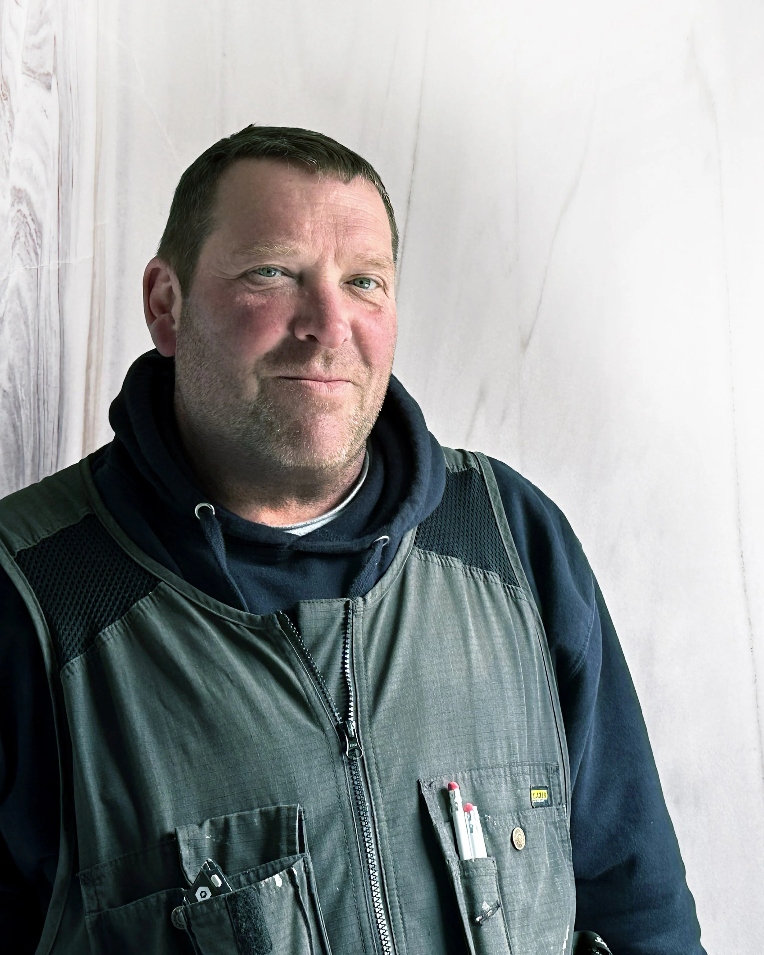 A man wearing a black hoodie and gray vest with pens in his vest pocket stands against a marble wall.